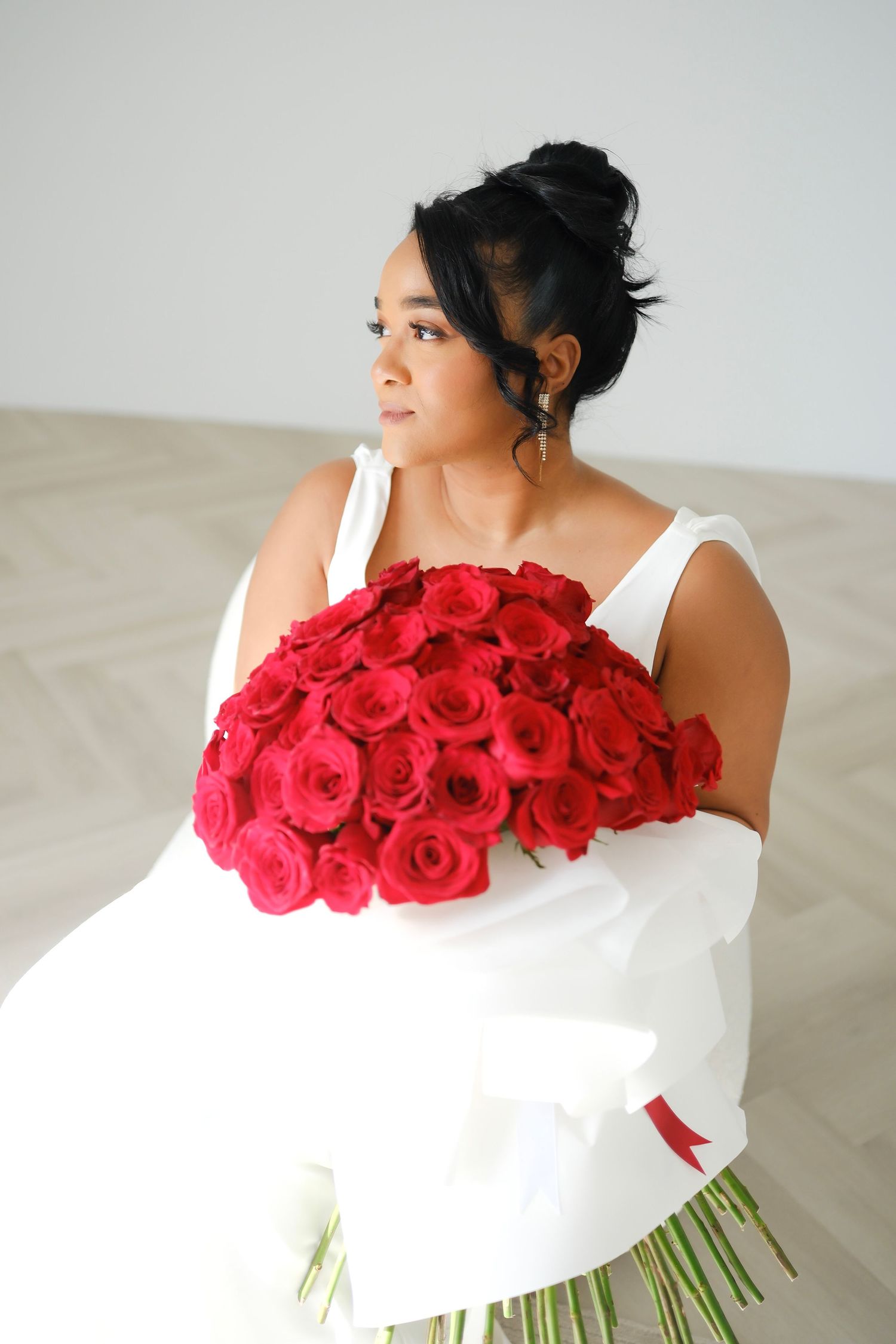 A person in a white off-shoulder top holds a stunning bouquet of vibrant red roses against a light background.