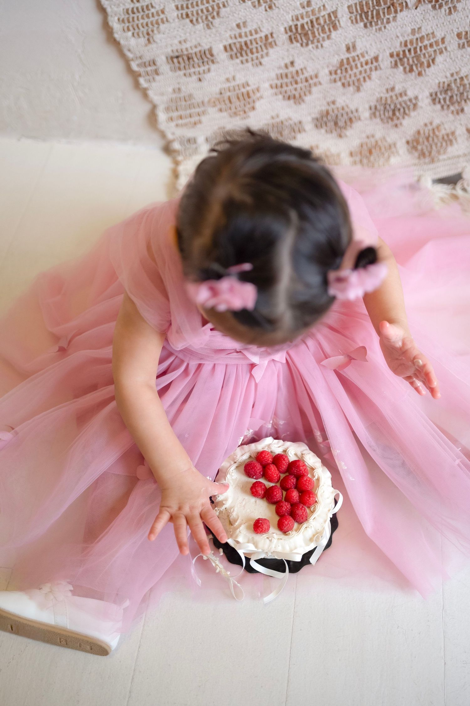 Toddler girl in pink tutu dress sitting on floor holding a white cake topped with fresh raspberries.