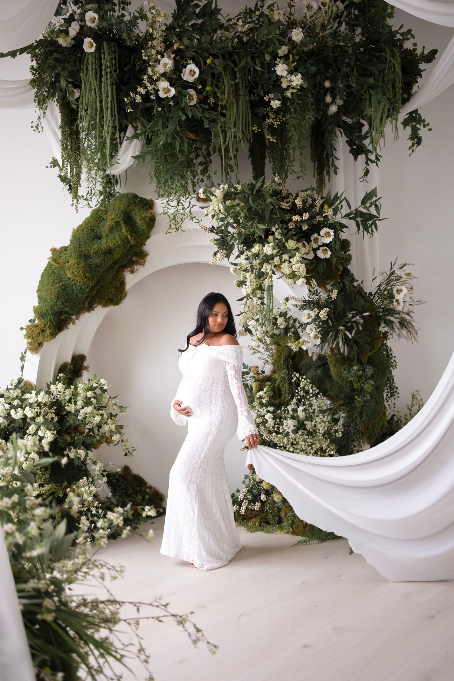 Pregnant woman in white off-shoulder gown poses among lush green floral arch with white blooms in elegant studio.