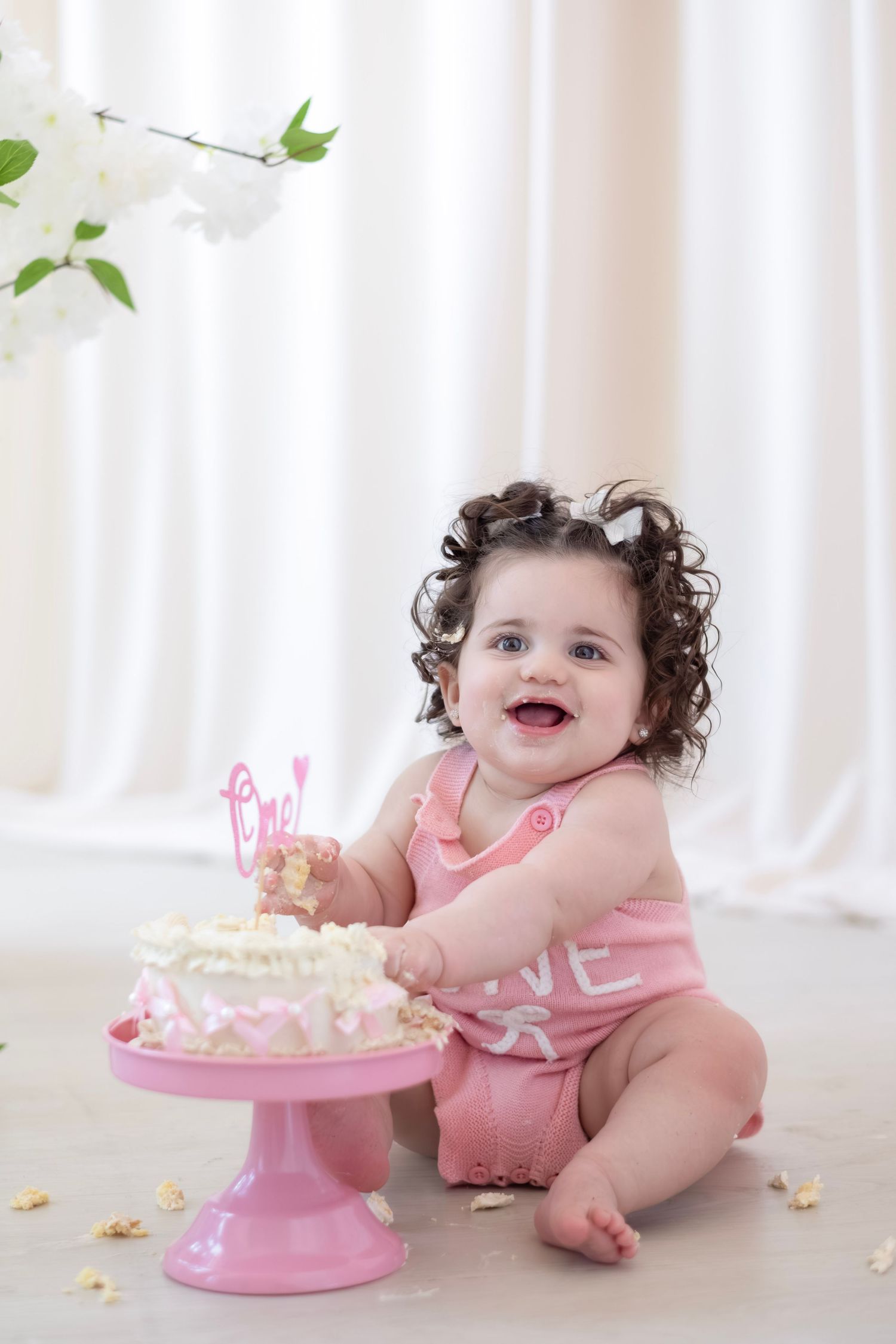 Smiling baby girl in pink romper enjoying her first birthday cake smash with white frosting on a pink cake stand.