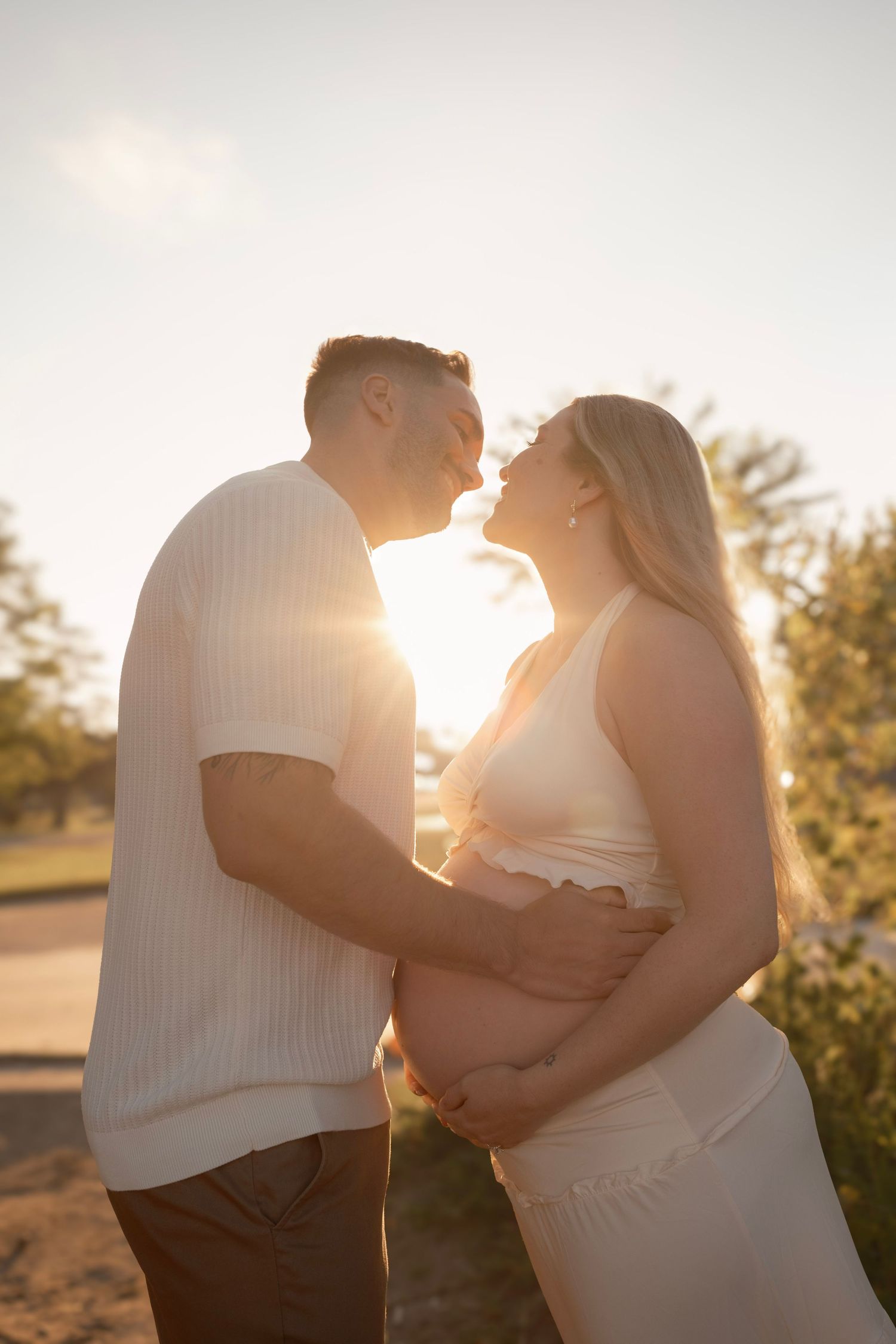 Couple sharing a tender moment at golden hour, pregnant woman cradling baby bump in soft backlit sunlight outdoors.