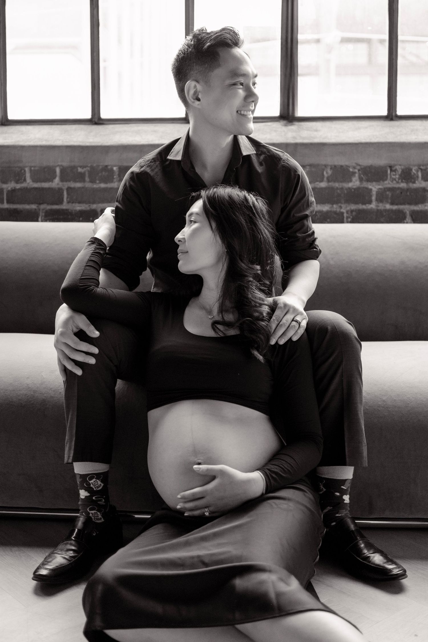 Black and white maternity photo of a pregnant woman and her partner sitting together on a couch, sharing a tender moment.
