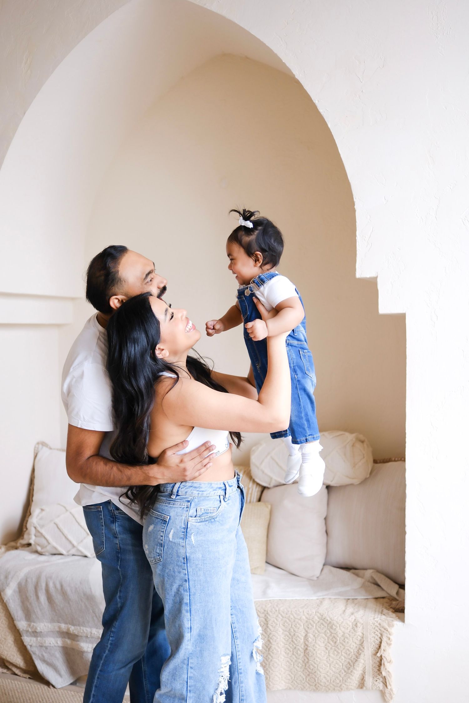 Family wearing denim and white tops plays together by an arched window alcove in natural lighting.