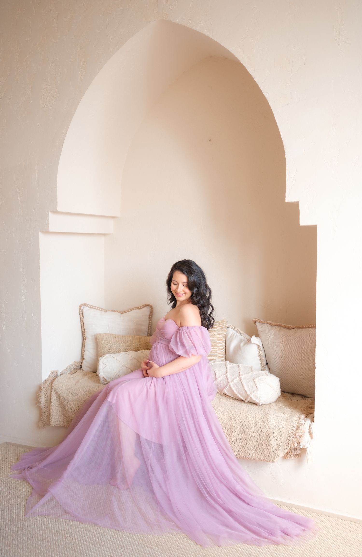 Figure in flowing lavender gown poses elegantly in arched alcove with cream pillows and soft lighting.