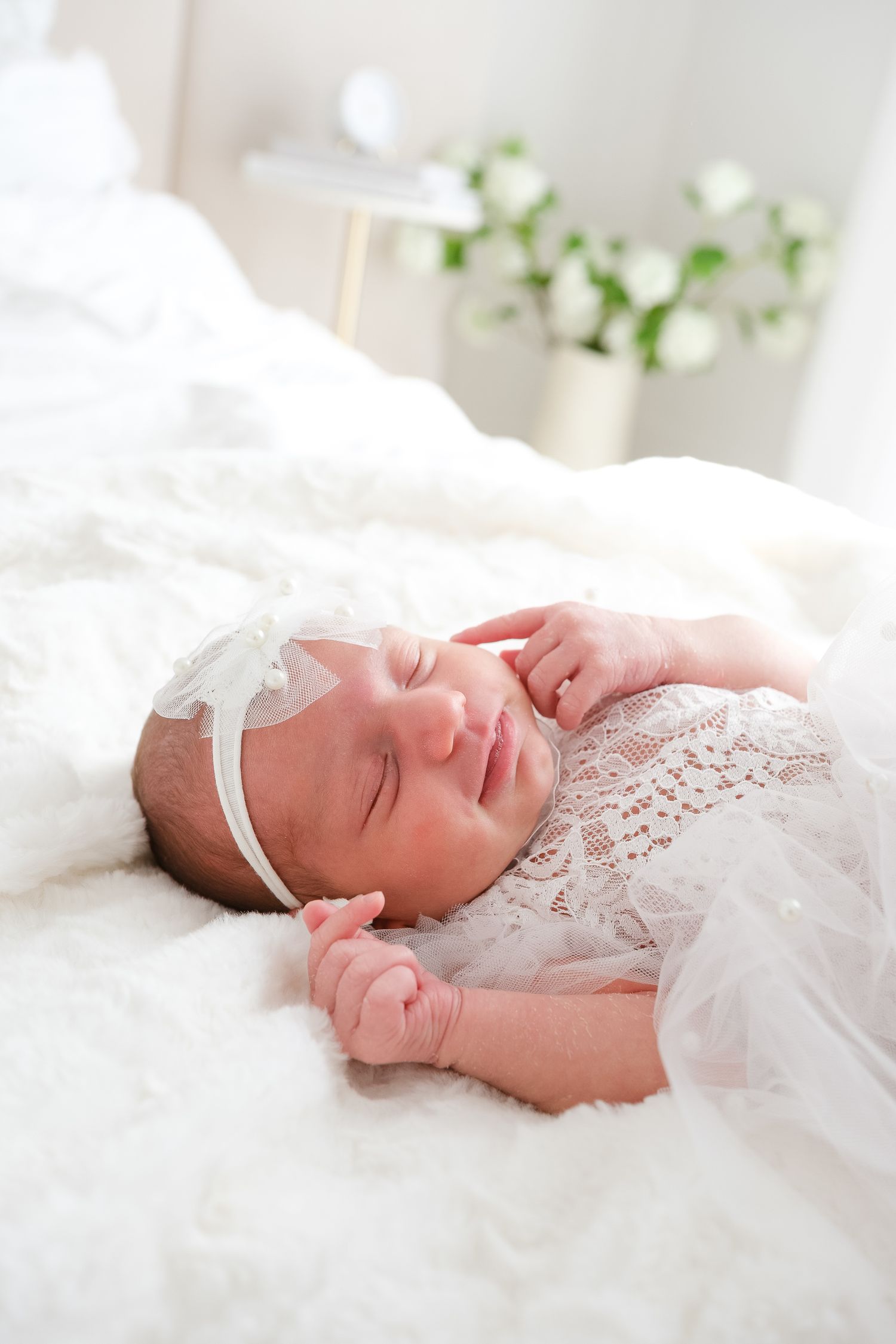A sleeping newborn baby wearing a white lace dress and headband lies peacefully on soft white bedding.