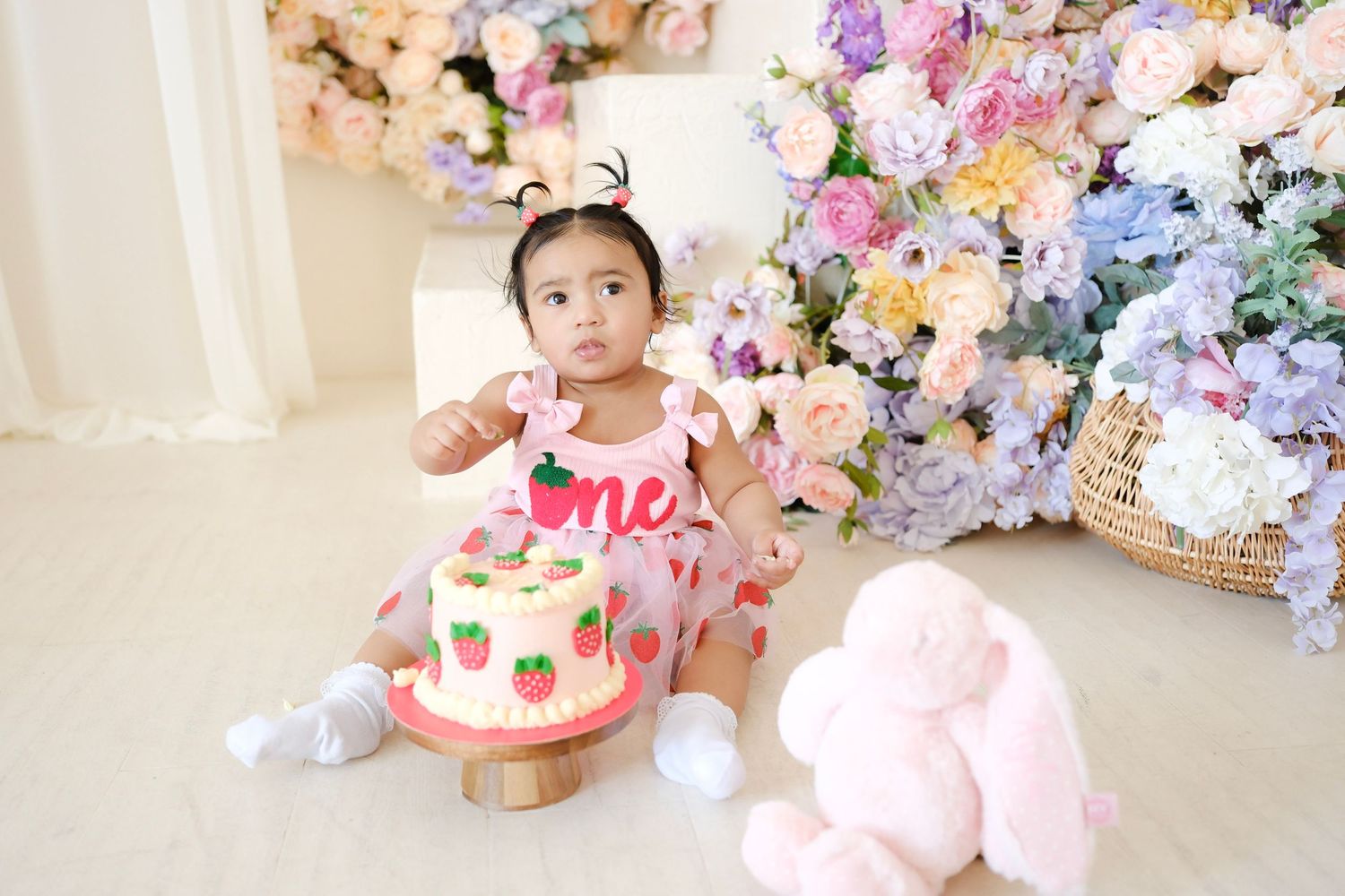 First birthday celebration setup with floral backdrop, pink cake on stand, and plush bunny toy decoration.