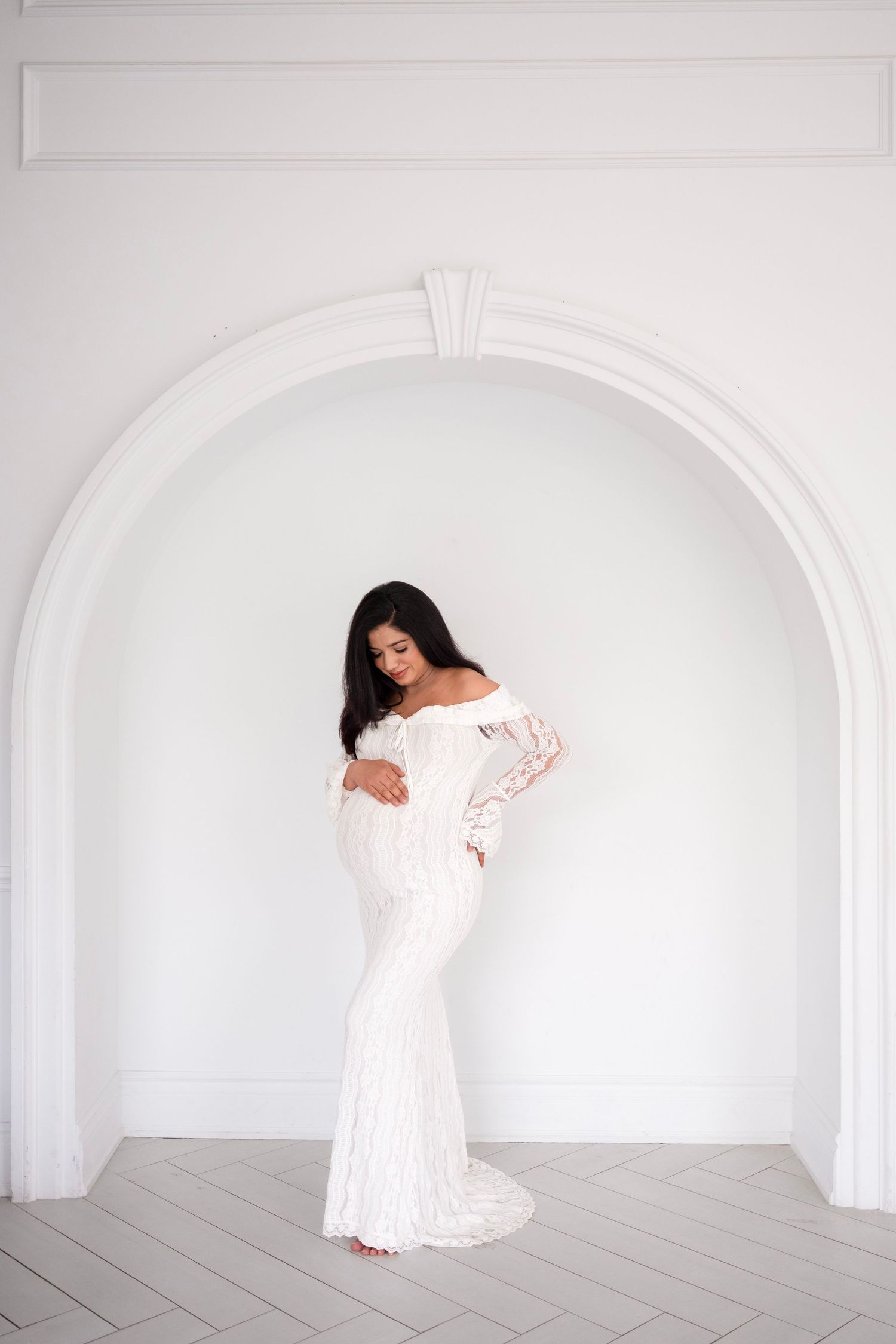 Pregnant woman in white lace off-shoulder gown poses gracefully in front of elegant white arched wall.