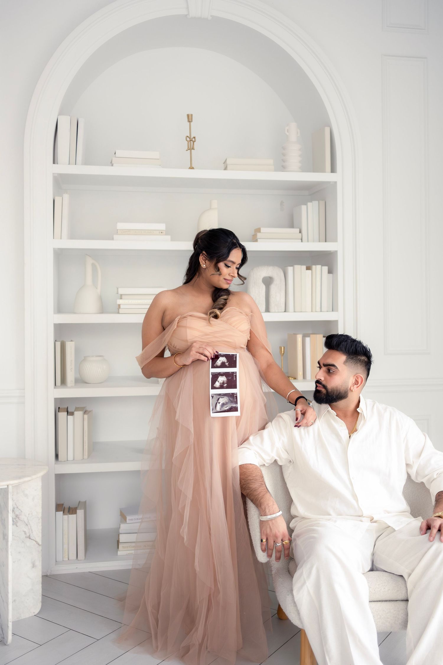 A maternity photography session in an all-white room with built-in shelves showing tender moments between a couple.