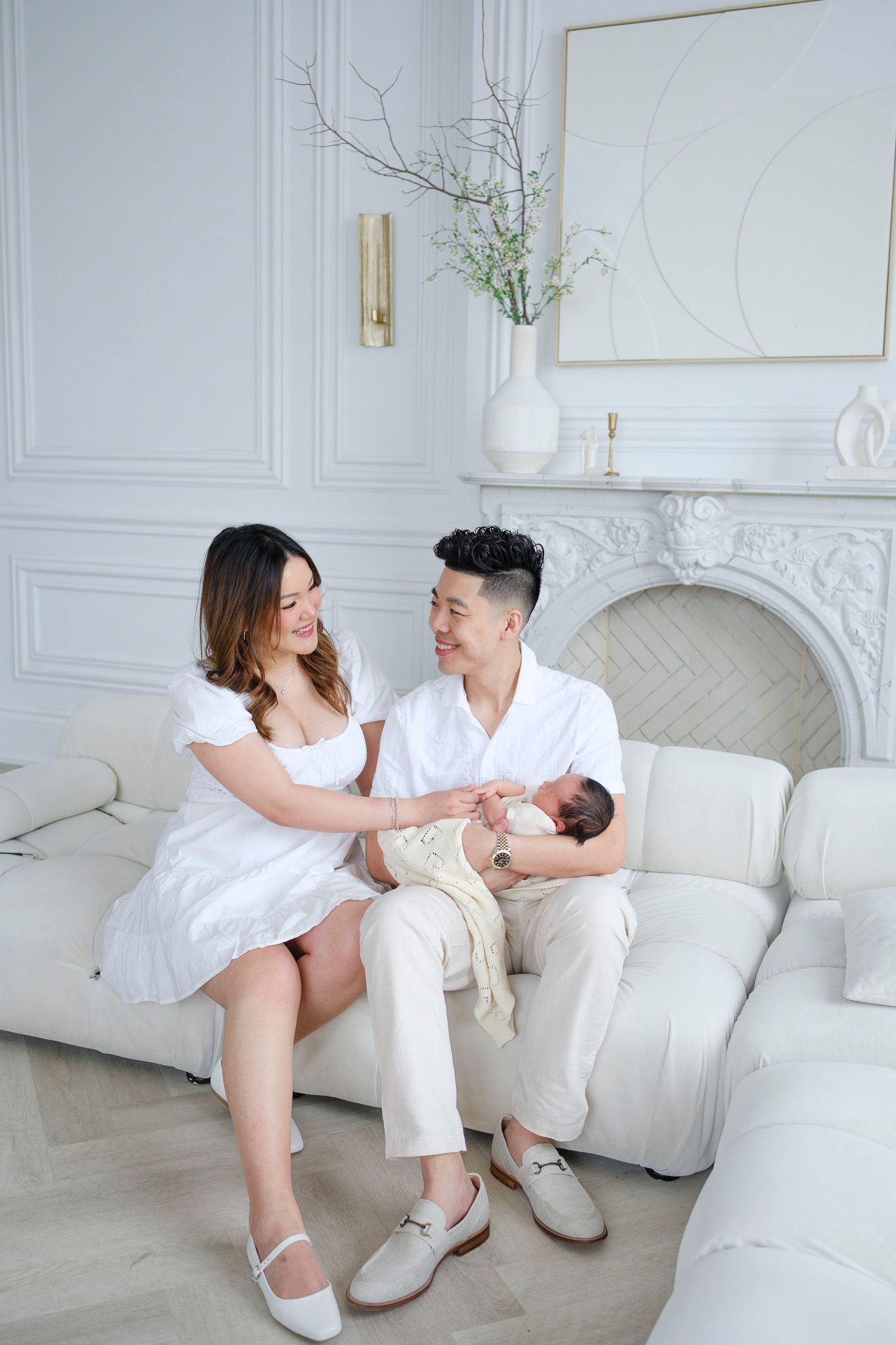 Couple in white outfits sitting together on plush sofa in elegant white room with ornate fireplace and floral decor.