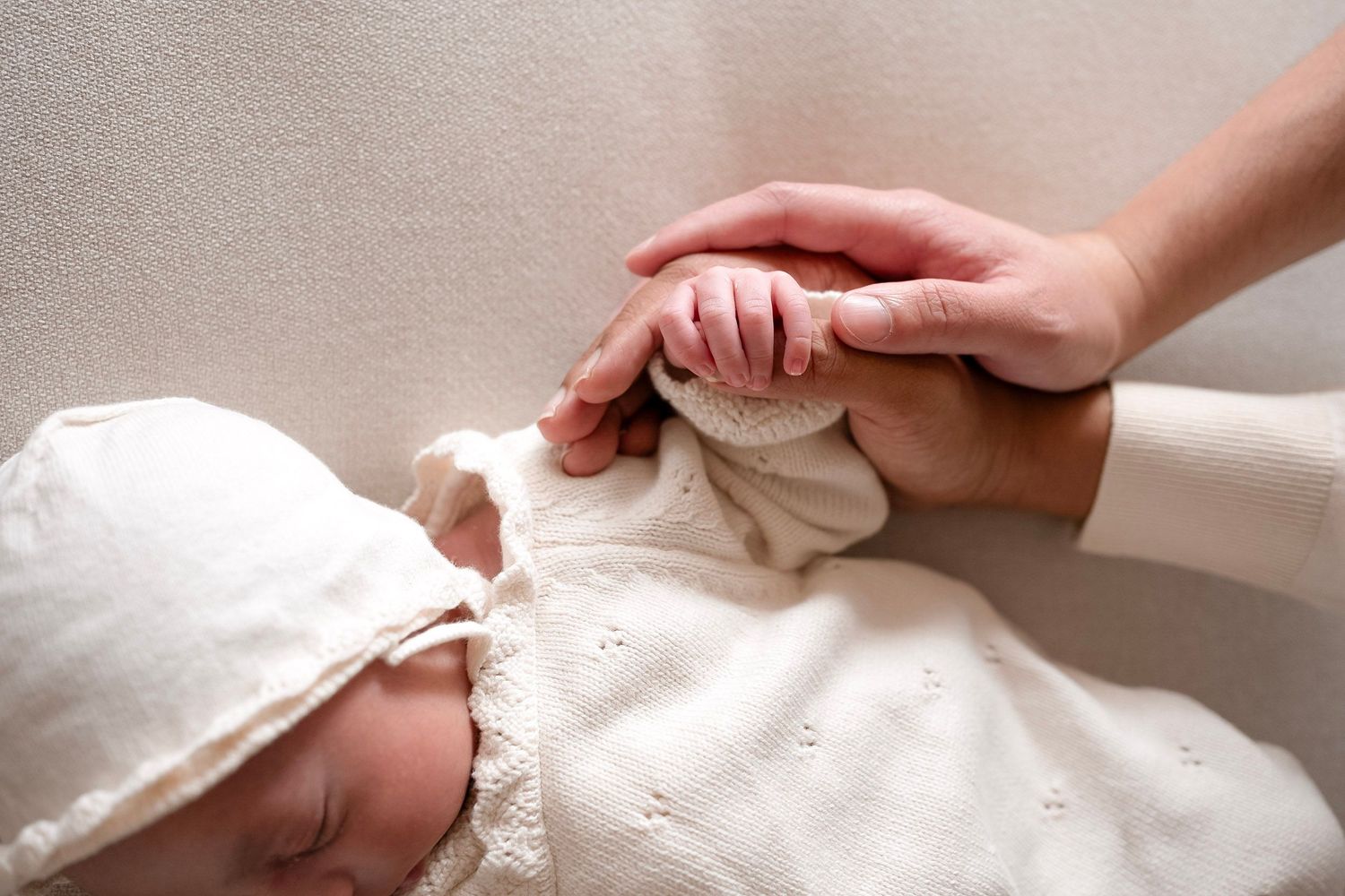 Sleeping newborn baby in white knit outfit and bonnet gently holding a parent's finger.