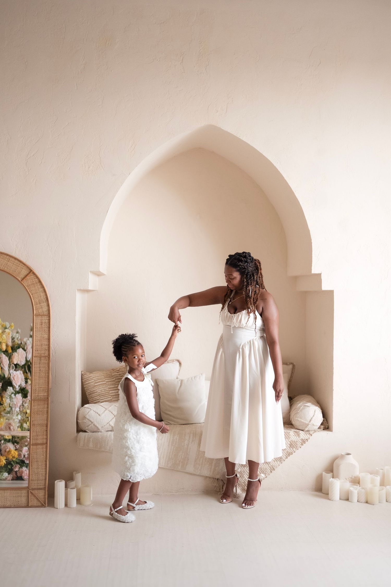A mother and daughter in matching white dresses hold hands and dance in an elegant cream-toned room with arched alcove.