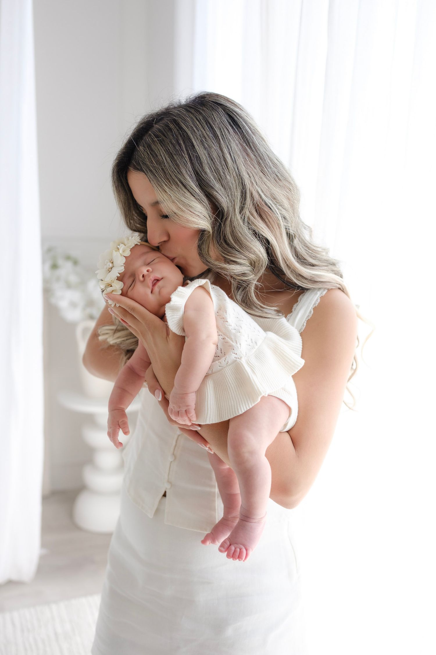A mother in a white dress tenderly kisses her newborn baby, both dressed in white, in a bright airy room.