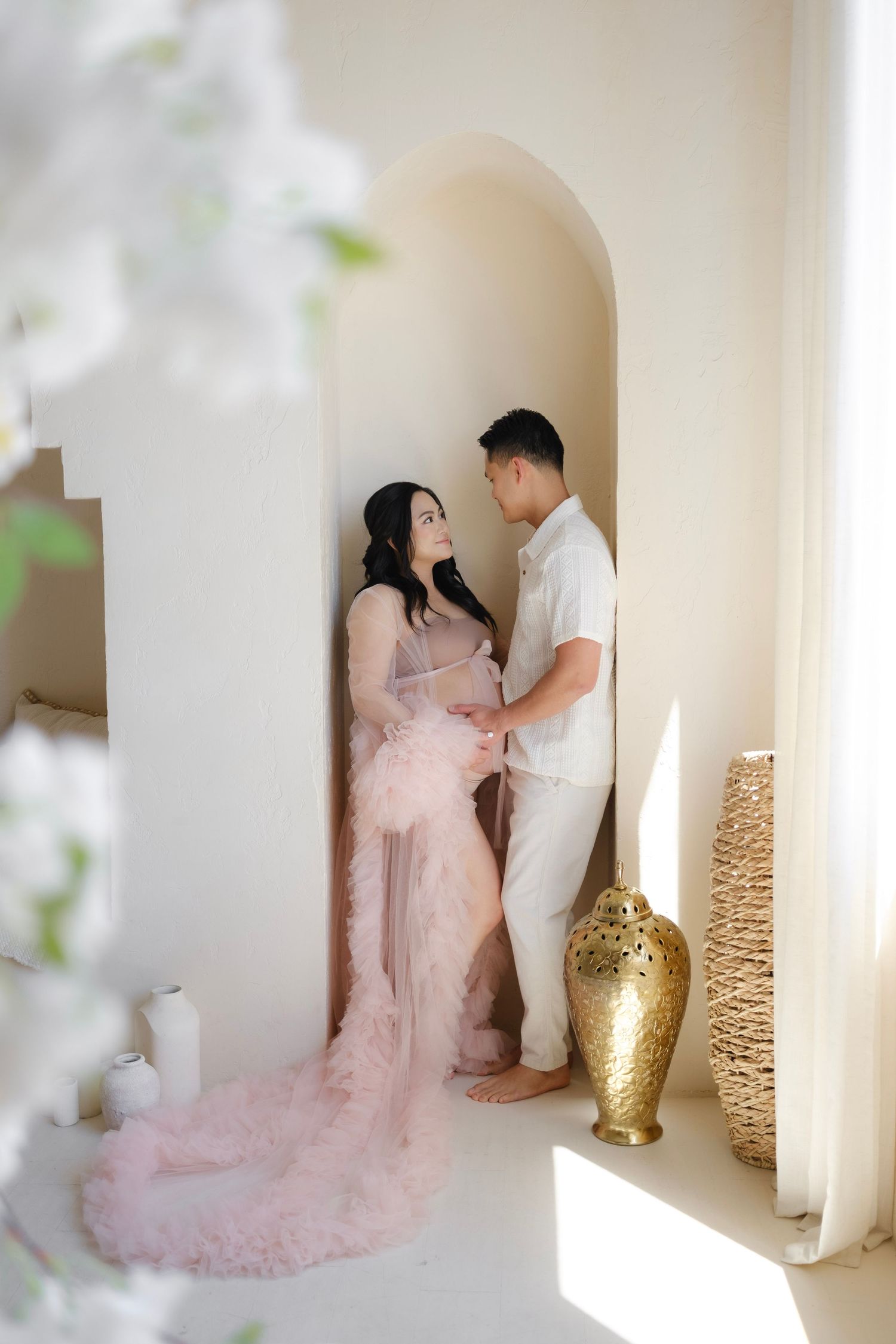 Pregnant couple sharing a tender moment in an elegant arched alcove, woman in pink tulle gown, soft white studio setting.