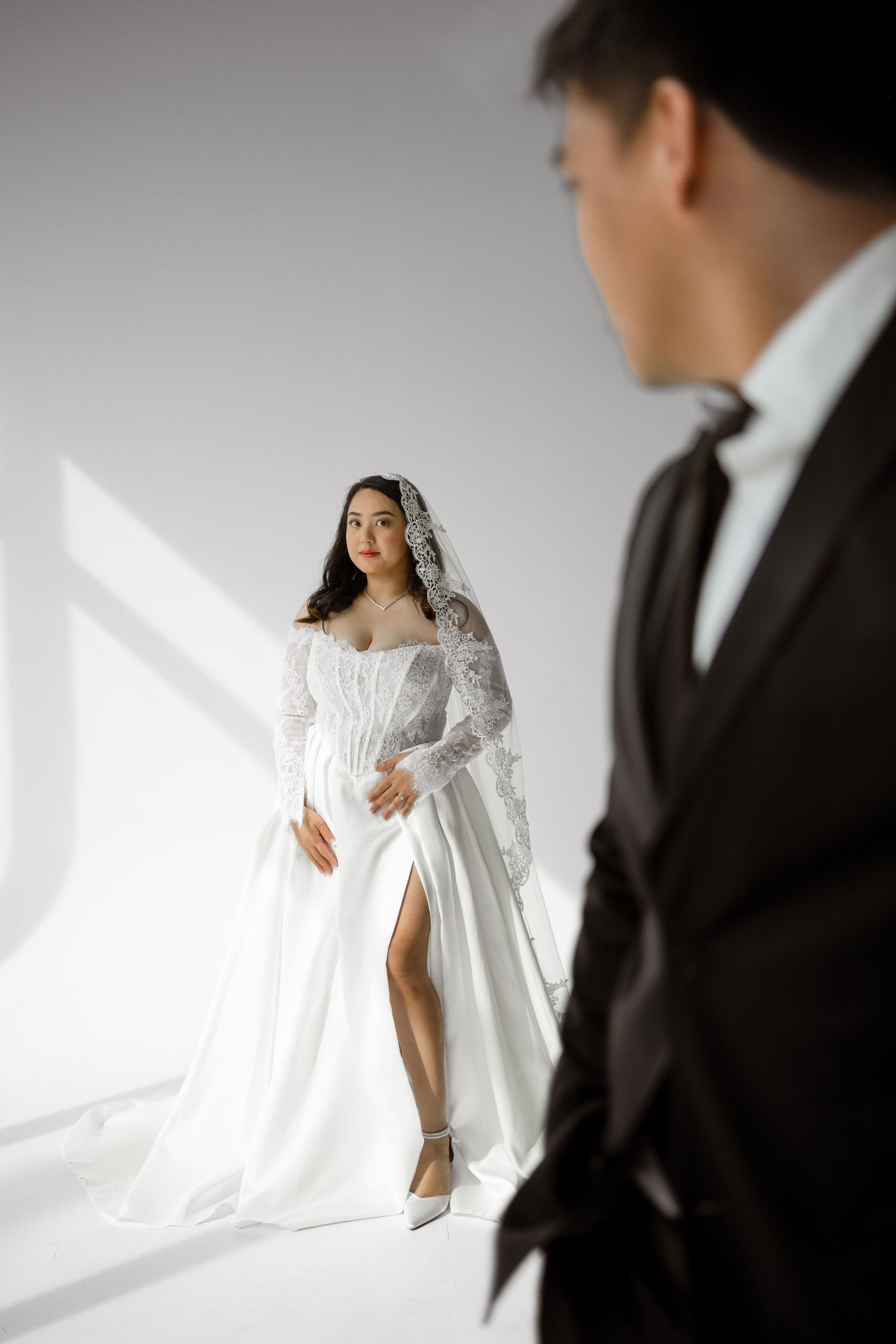 Elegant bride in white lace gown with veil poses as groom stands in foreground in black suit.