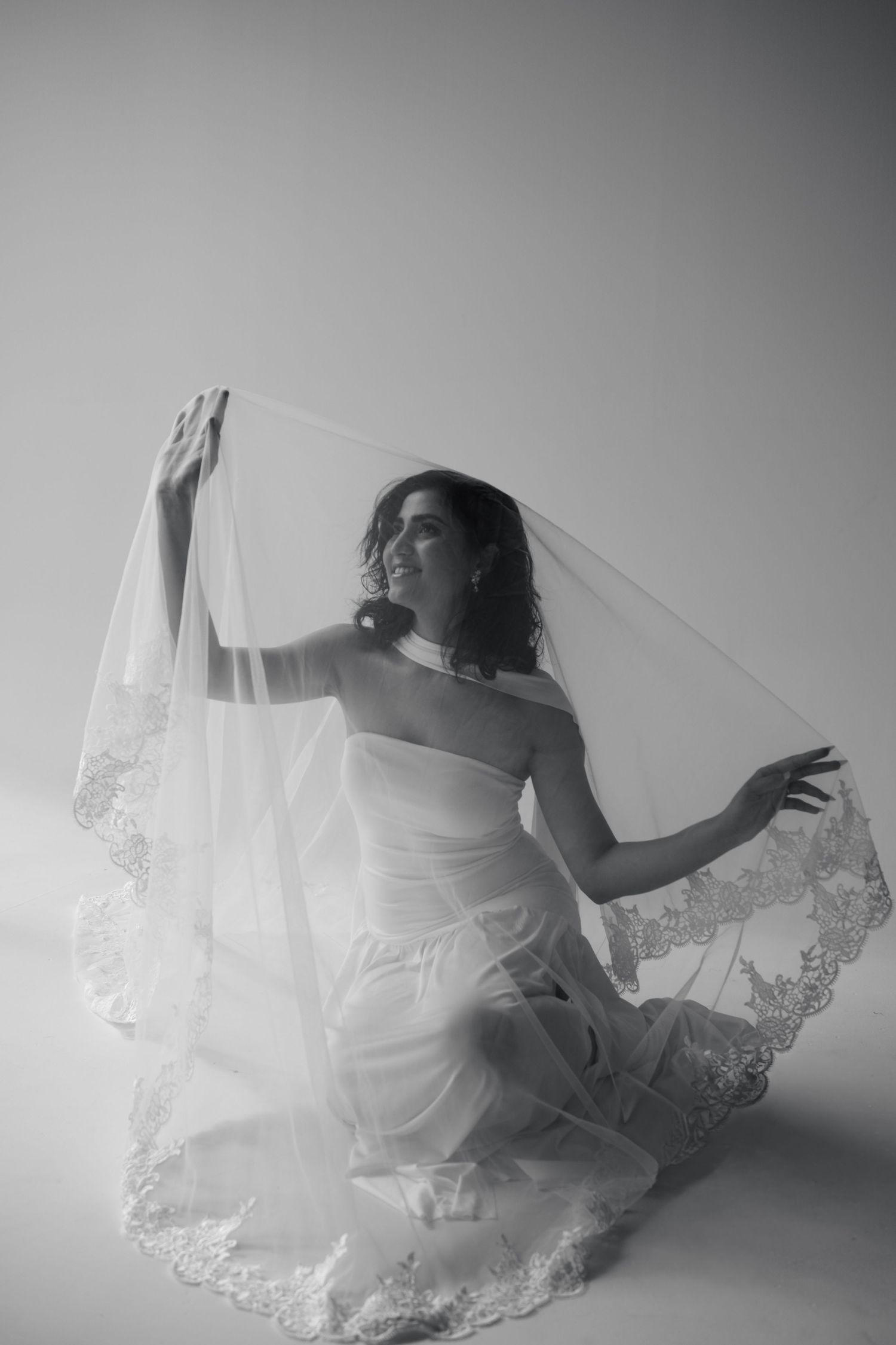 Black and white photo of a bride in a strapless gown lifting her lace-trimmed veil gracefully.