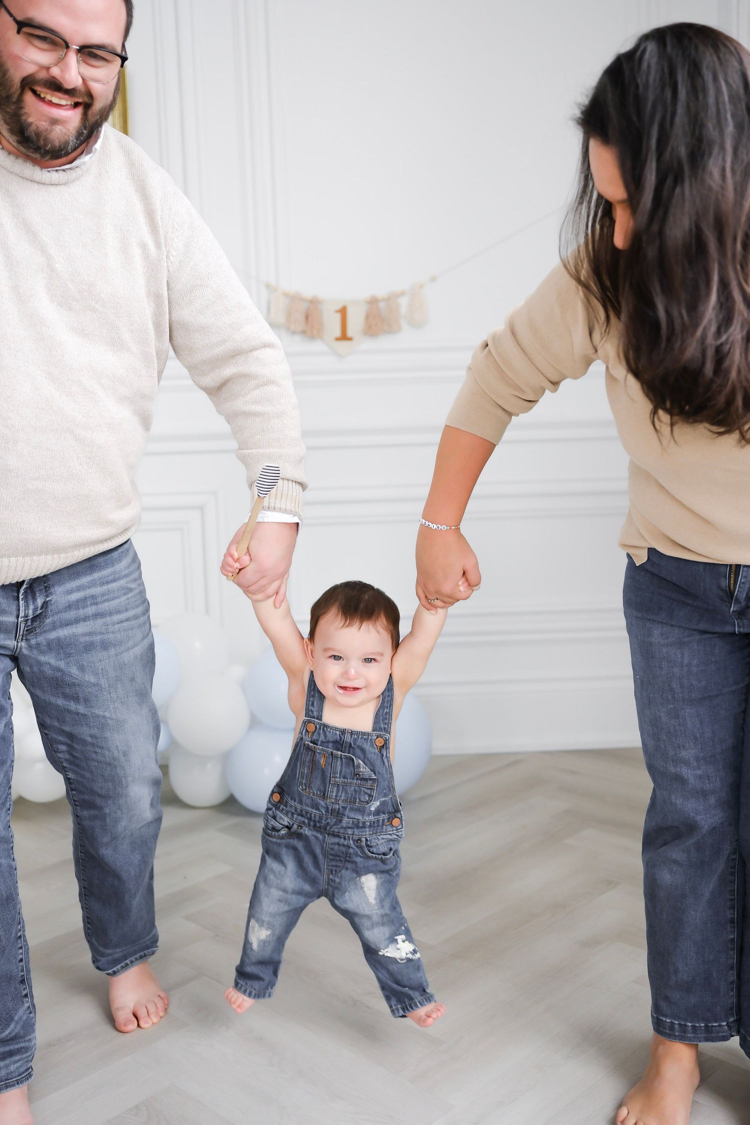 Happy baby in denim overalls takes first steps holding parents hands at first birthday party.
