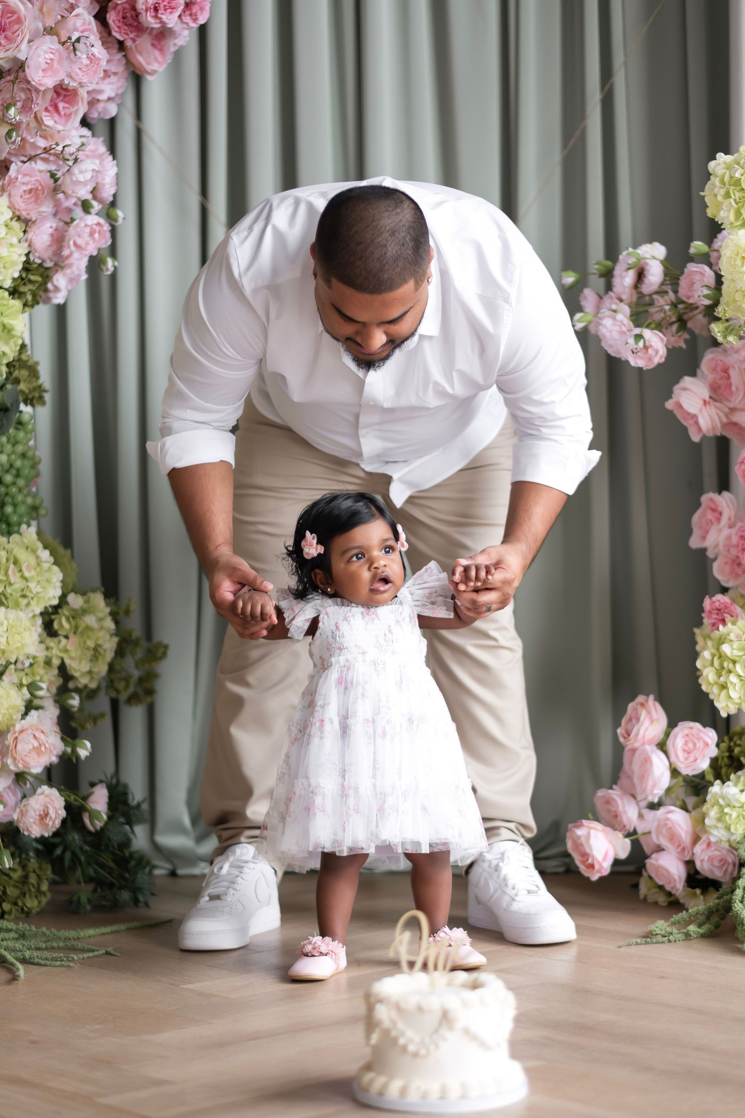 Father helps toddler girl in white dress stand near first birthday cake surrounded by pink floral decorations.
