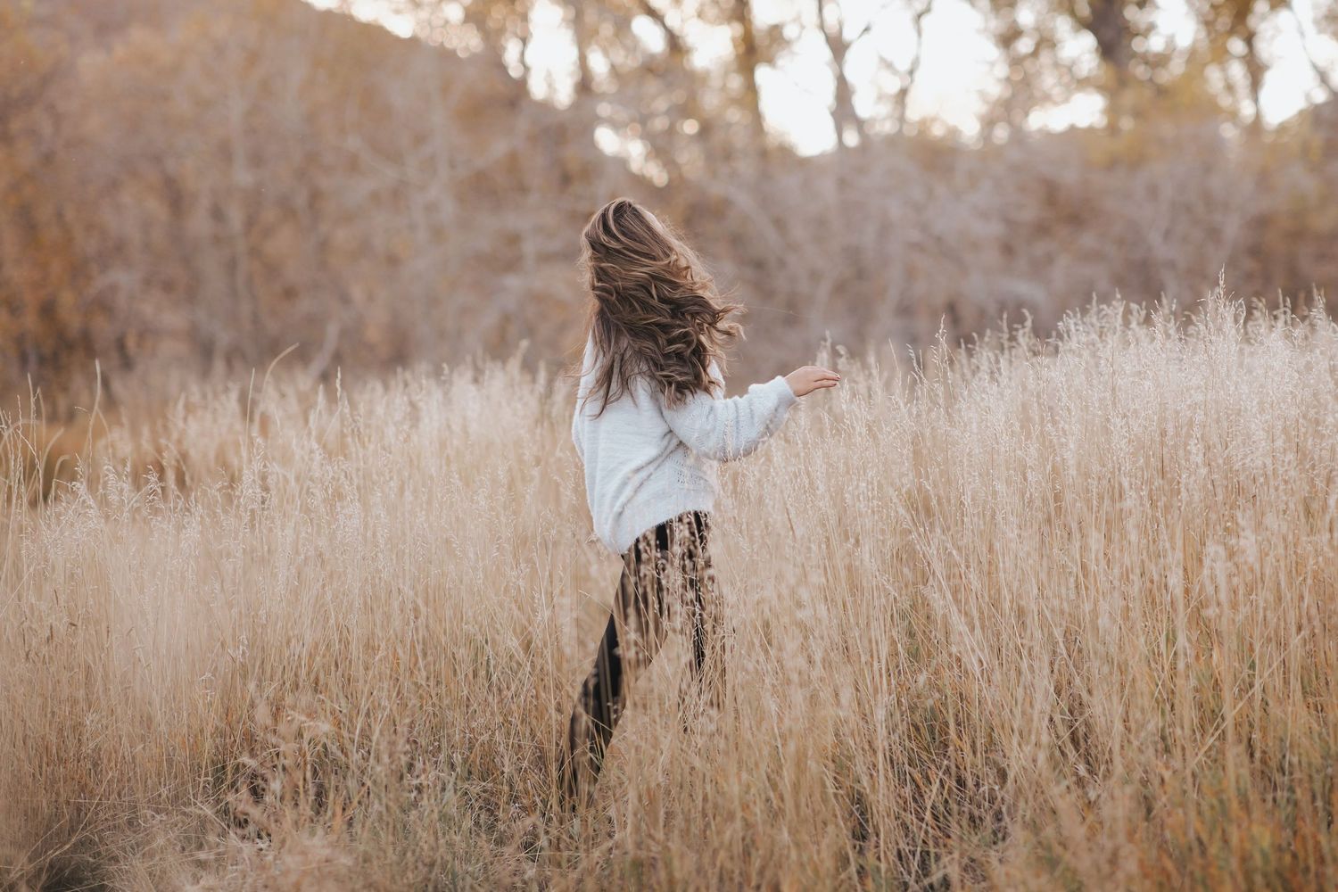 A young girl with long, flowing hair walks through a golden field of tall grass, her face turned away as she moves freely in the warm, autum