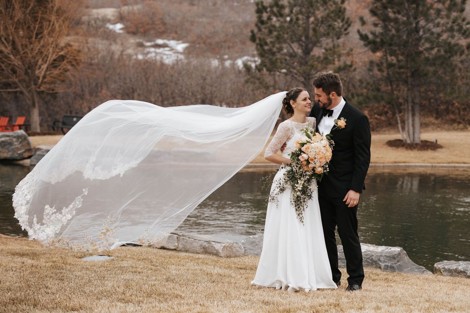 Bride and groom smile by a river as the bride’s veil flows in the wind, capturing a romantic wedding moment.