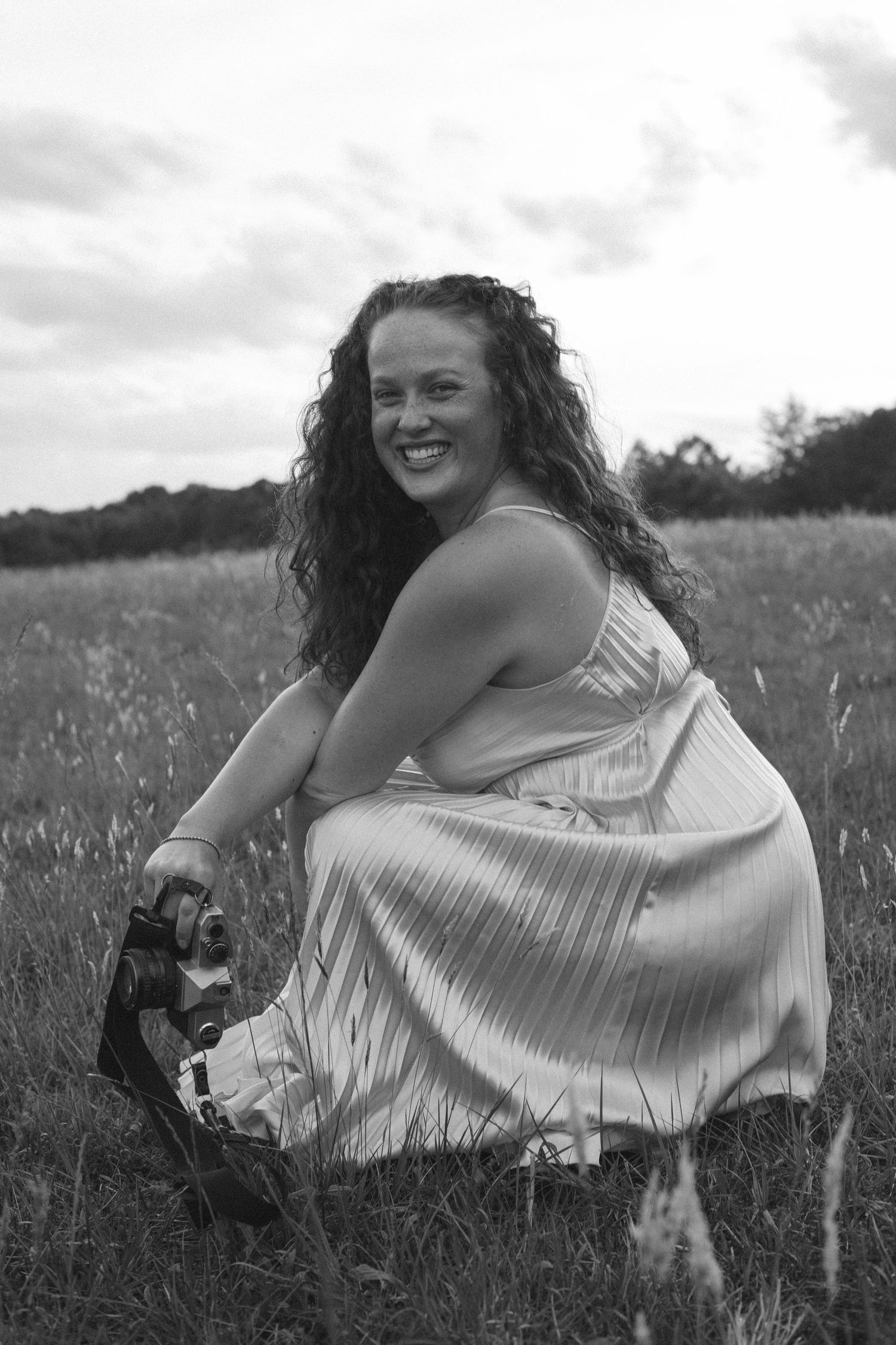 Black and white photo of a smiling woman with curly hair crouching in a grassy field wearing a flowy dress.