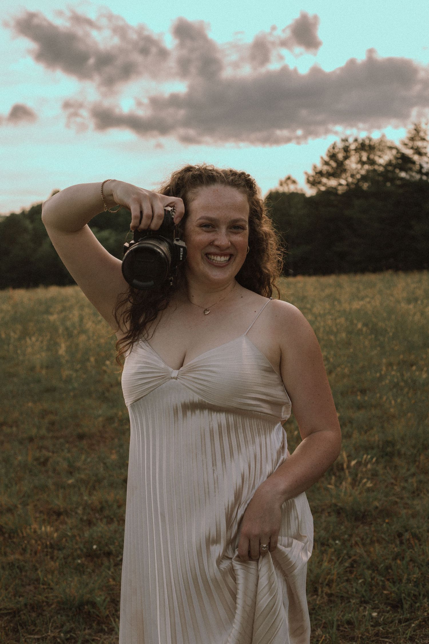 A smiling woman in a white slip dress holds a camera up near her face in a golden hour field.