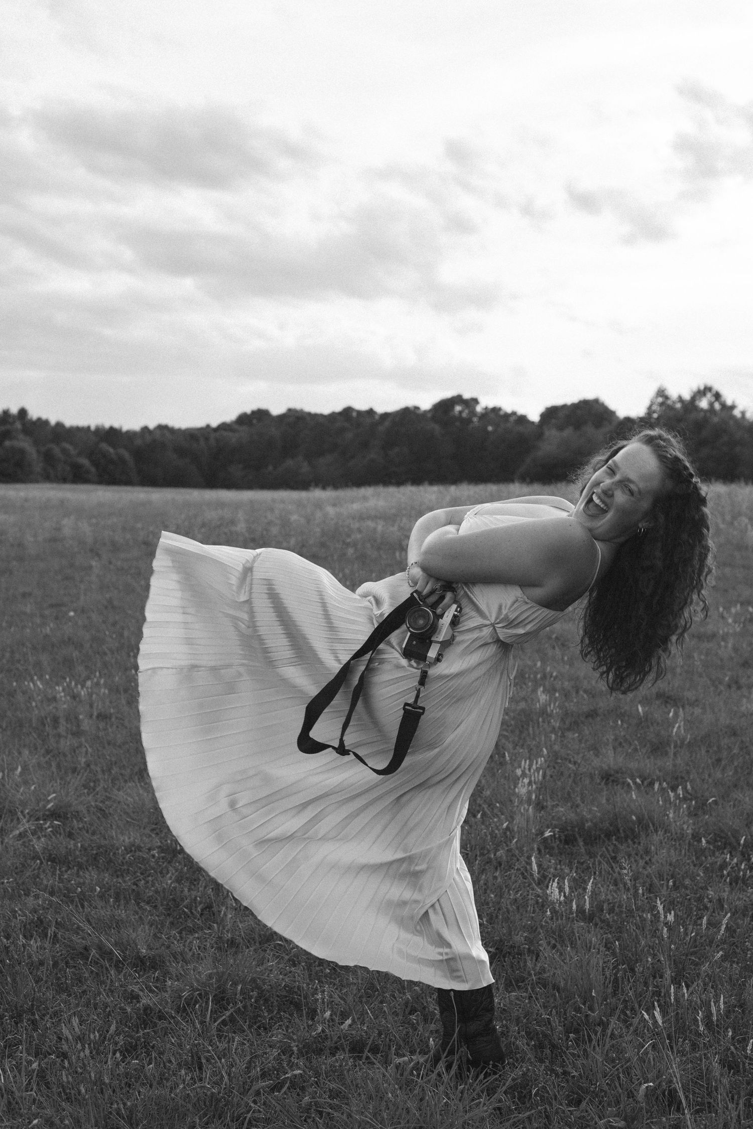 Black and white photo of a joyful woman in a flowing white dress dancing in an open field on a cloudy day.
