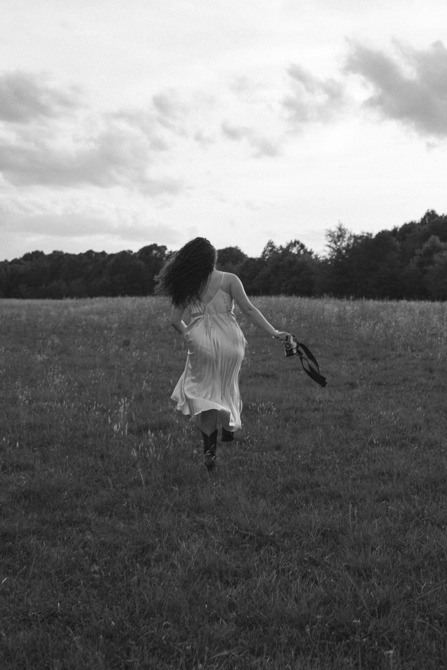 Black and white photo of a woman in a white dress running through a field, holding a hat, with trees in the background.