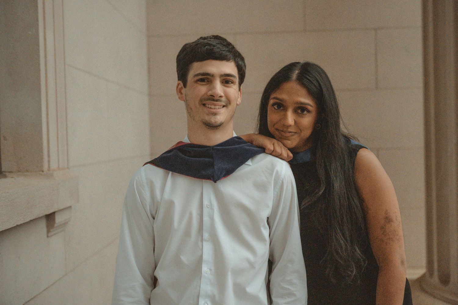 A young man in graduation attire poses with a woman in a black dress near stone columns.