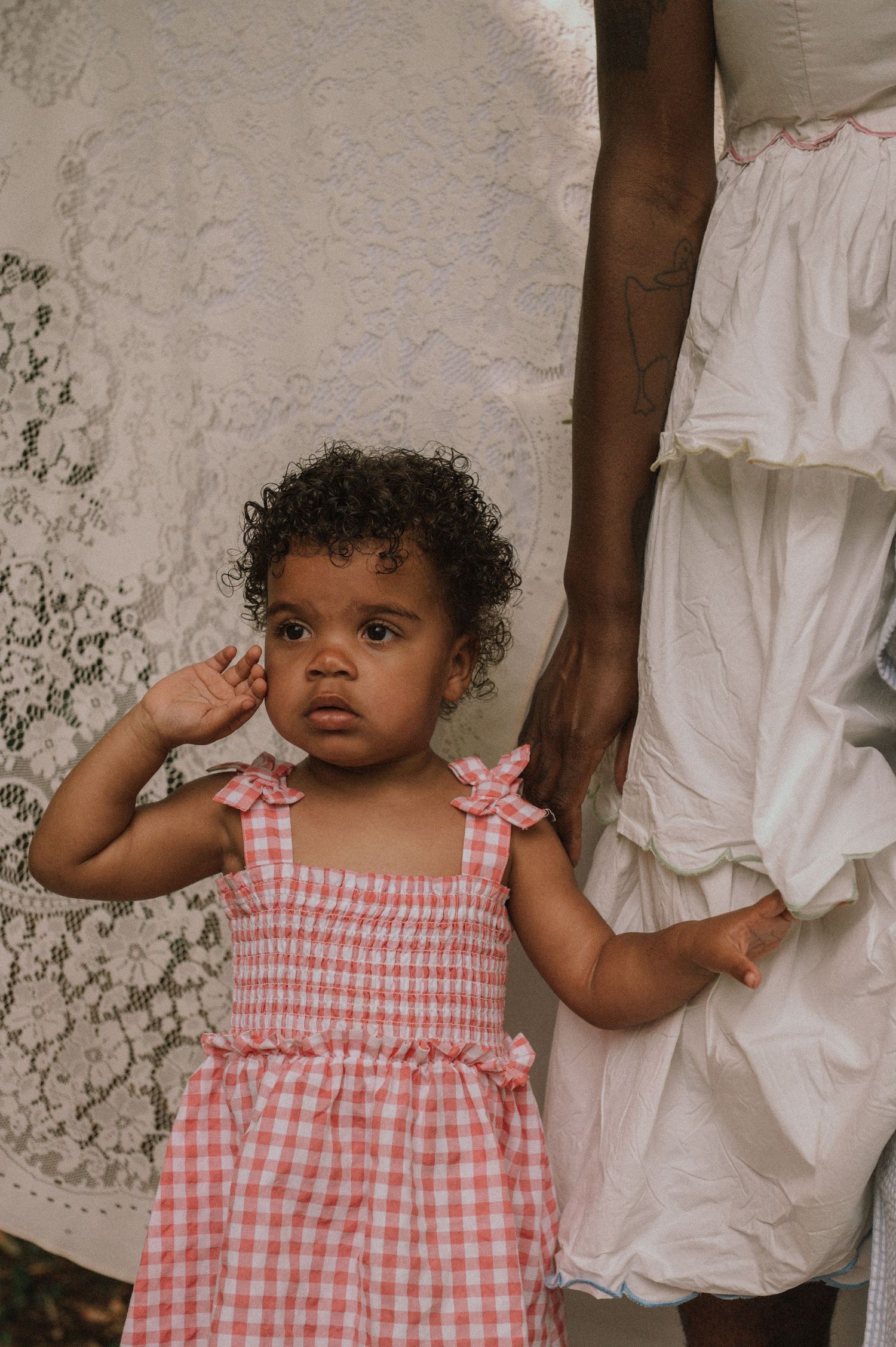 An adorable toddler in a pink gingham dress stands beside a person in a white lace gown outdoors.