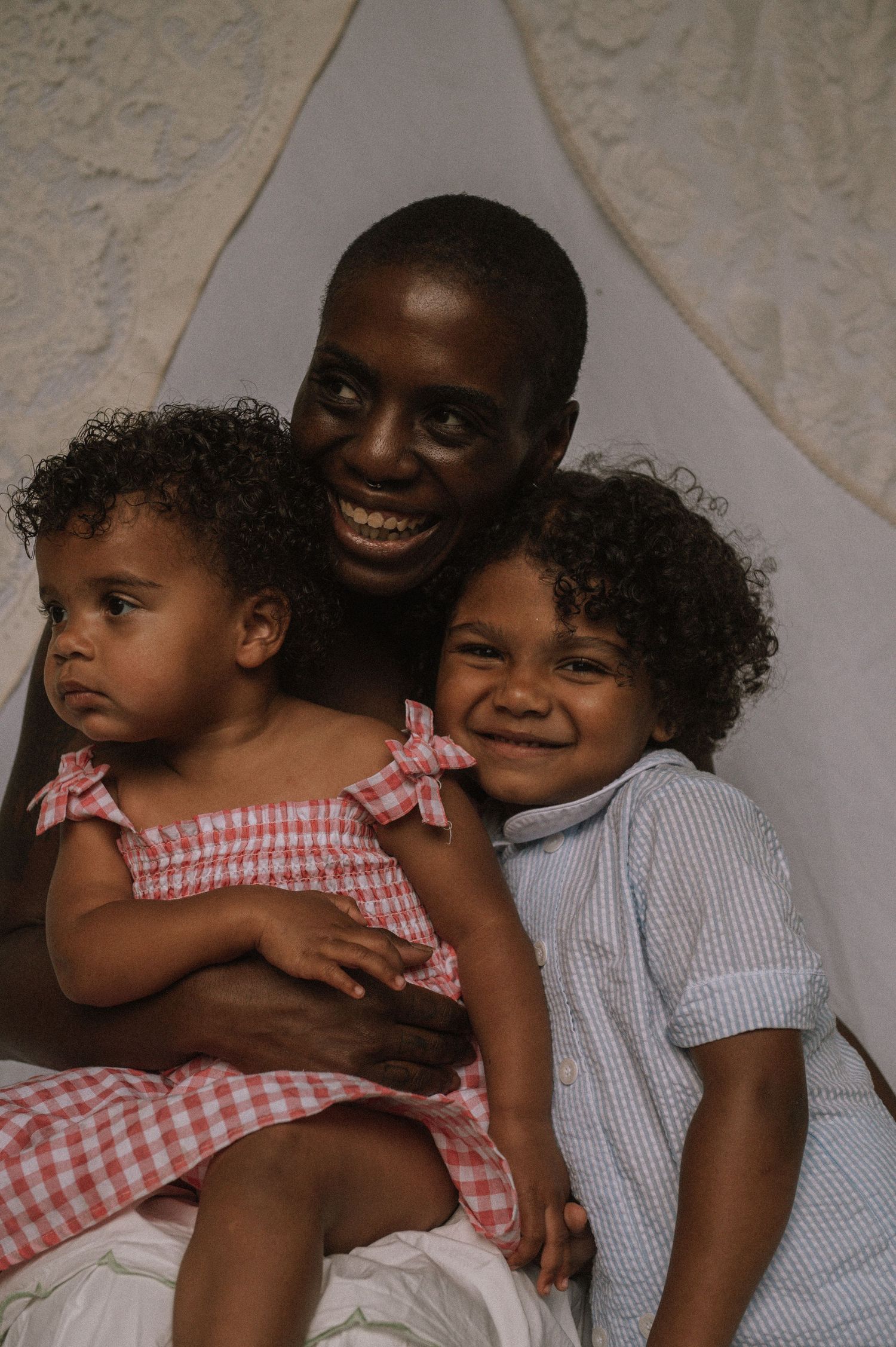 Smiling parent holding two young curly-haired children inside a white canopy tent, wearing gingham and striped outfits.