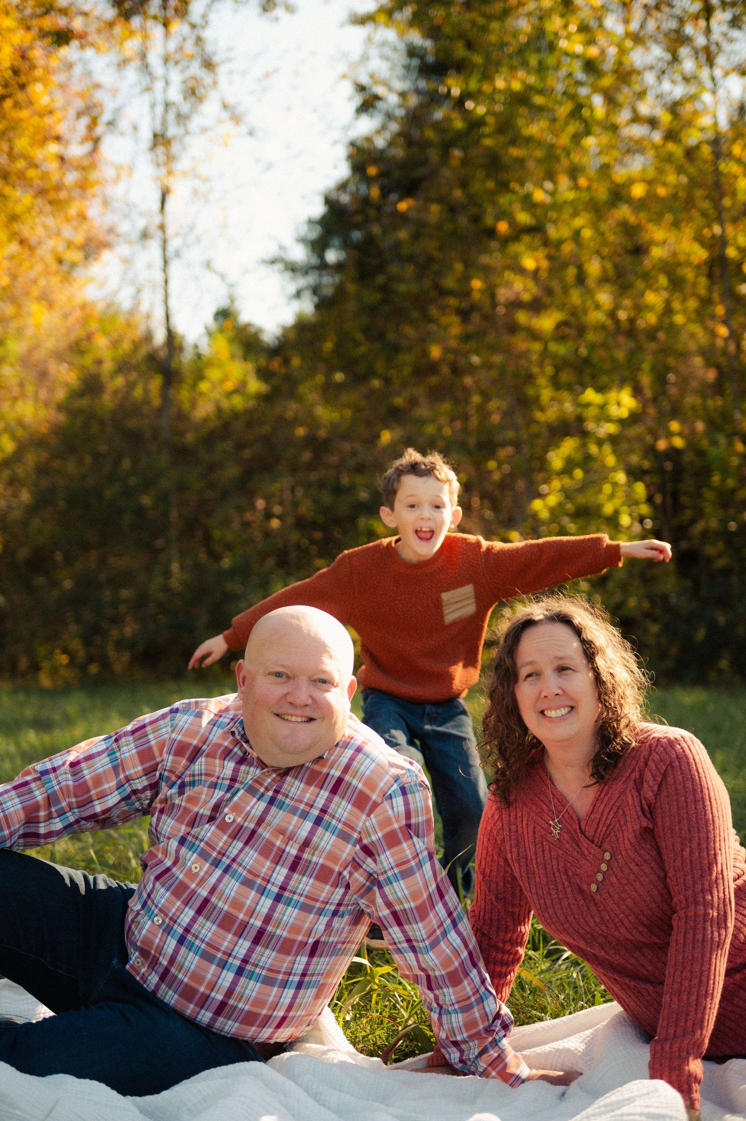 Family of three relaxing on a blanket outdoors with an energetic young boy jumping behind his smiling parents in fall.