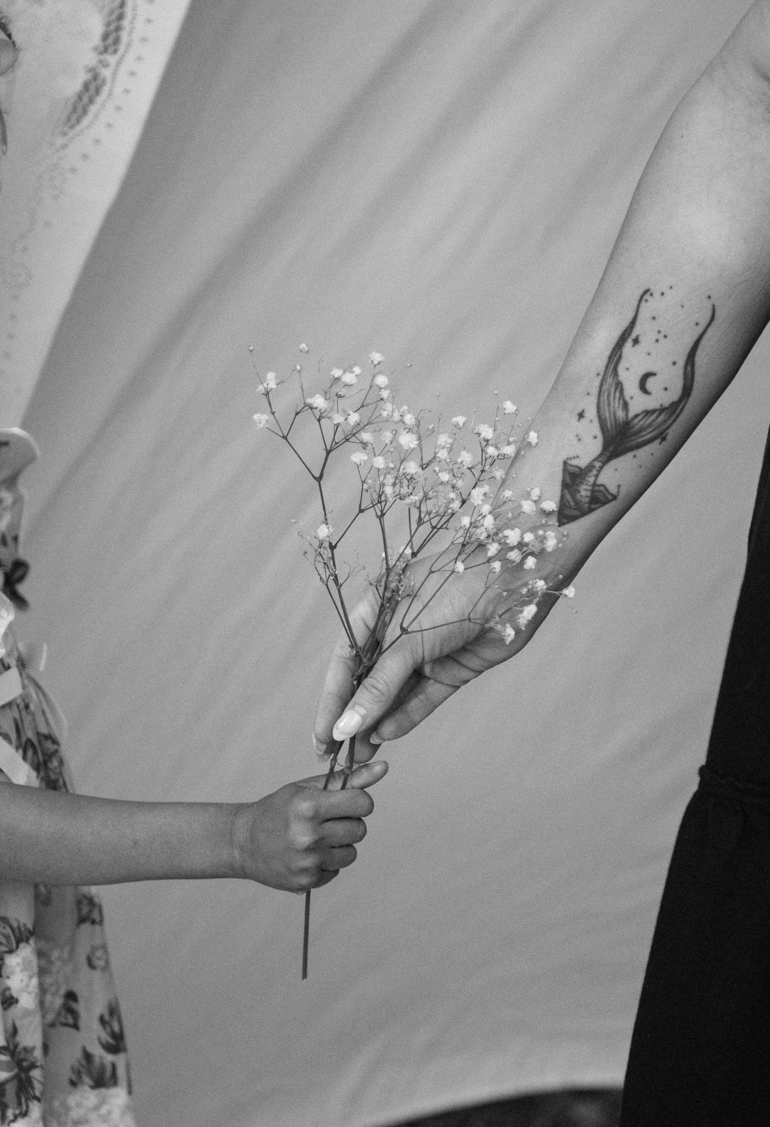 A child hands baby's breath flowers to a tattooed adult arm in a black and white artistic photo.