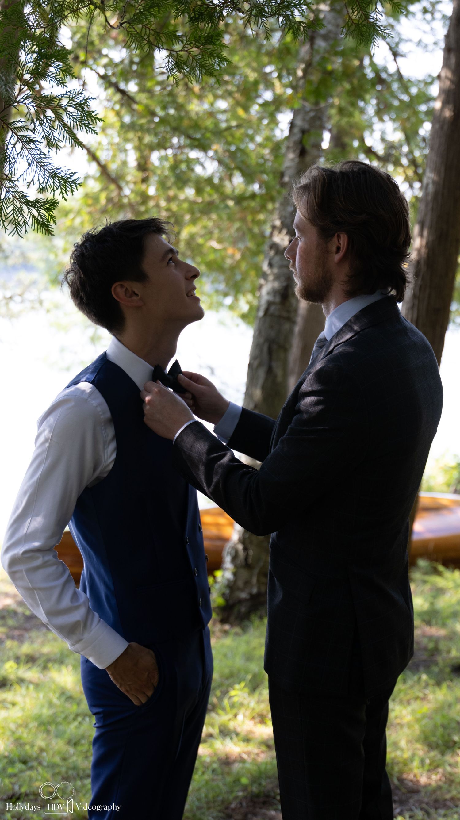 Two people in formal attire stand among trees by a lake while adjusting a tie.
