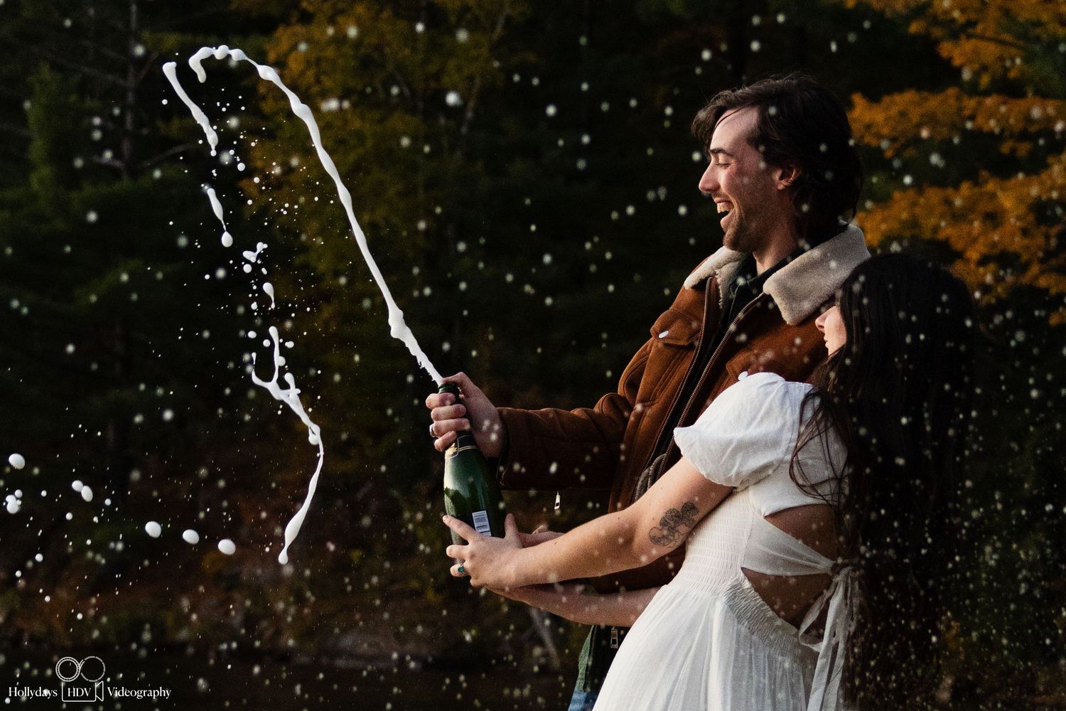 Couple celebrating with champagne spray during outdoor engagement photo session, captured in dramatic lighting with fall foliage background.