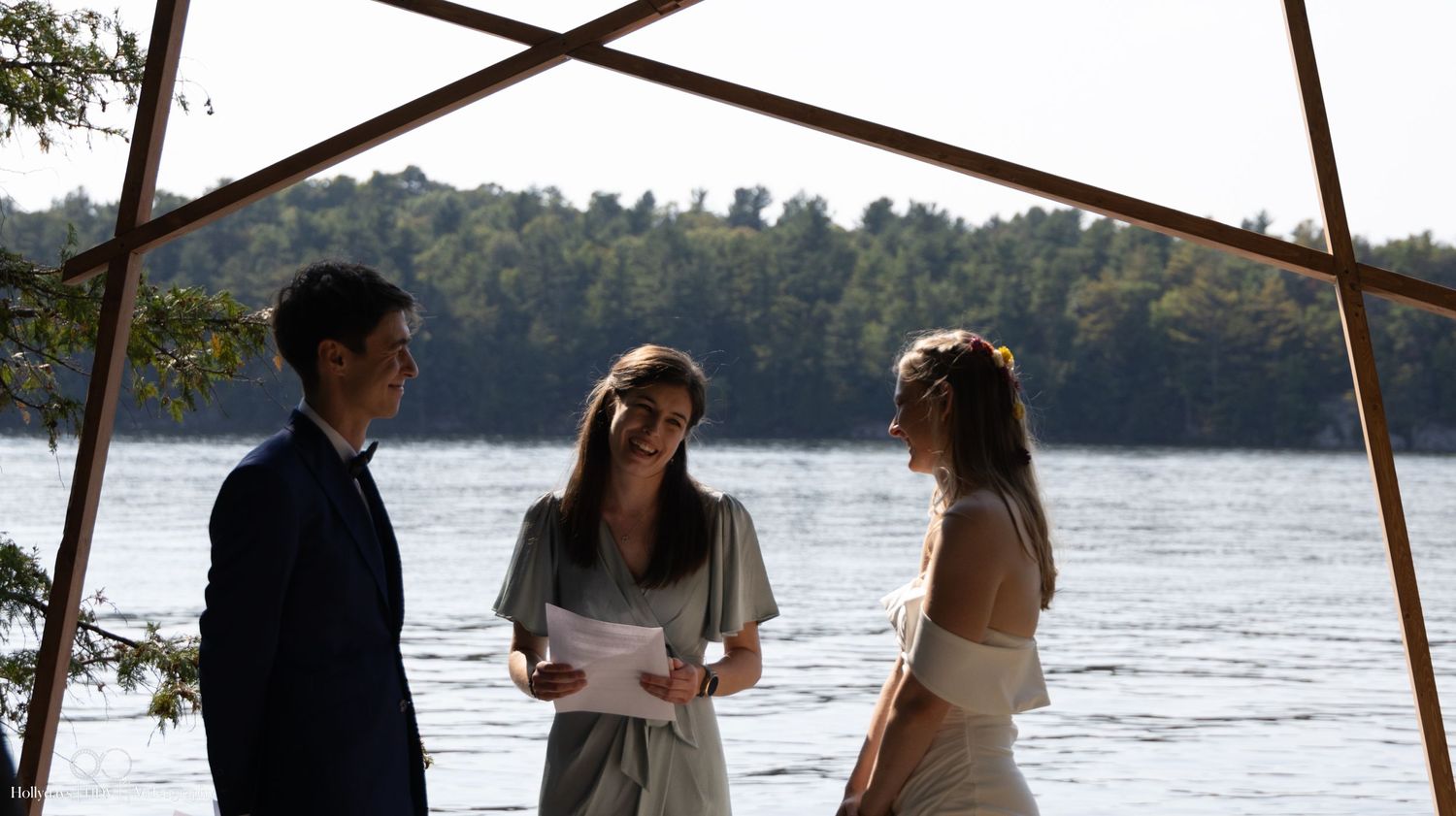 A wedding ceremony takes place under a wooden frame structure by a peaceful lakeside.
