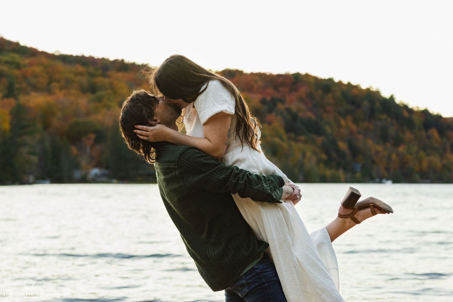 Romantic couple embraces by a scenic autumn lake with colorful foliage-covered hills in the background.