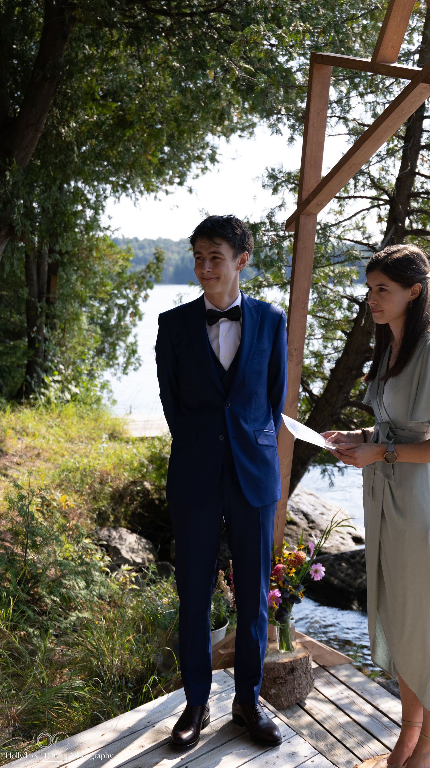 A wedding ceremony participant stands in a blue suit on a deck overlooking a lake.