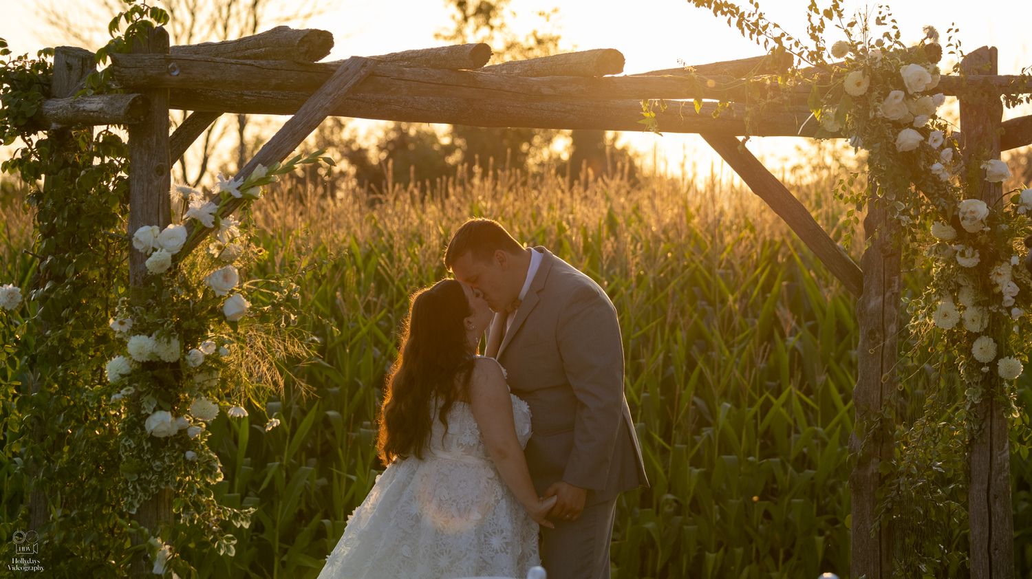 A romantic golden hour wedding moment under a rustic wooden arch adorned with white flowers in a field at sunset.