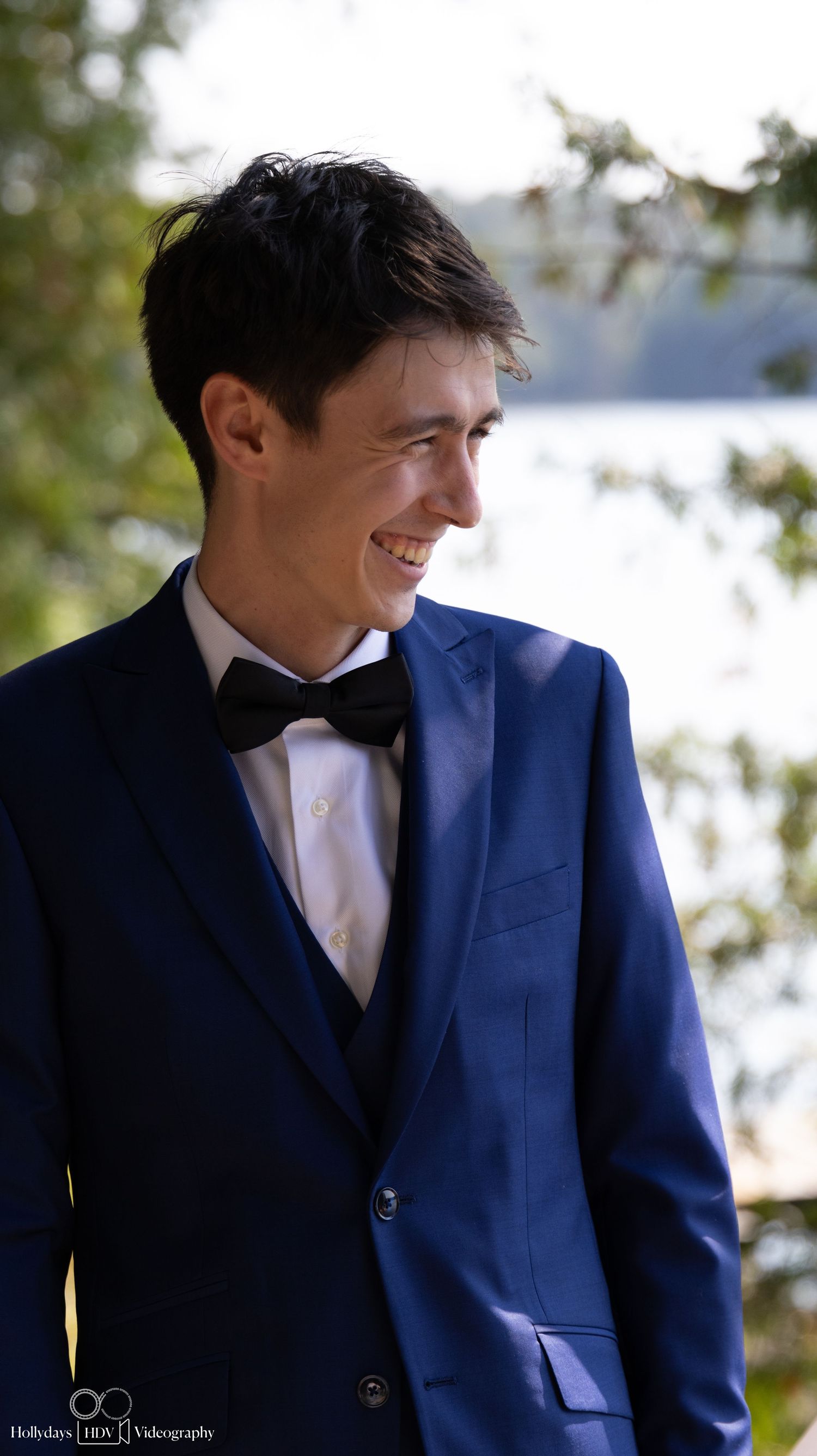 A person in a royal blue suit and bow tie smiles while standing outdoors near a lake.