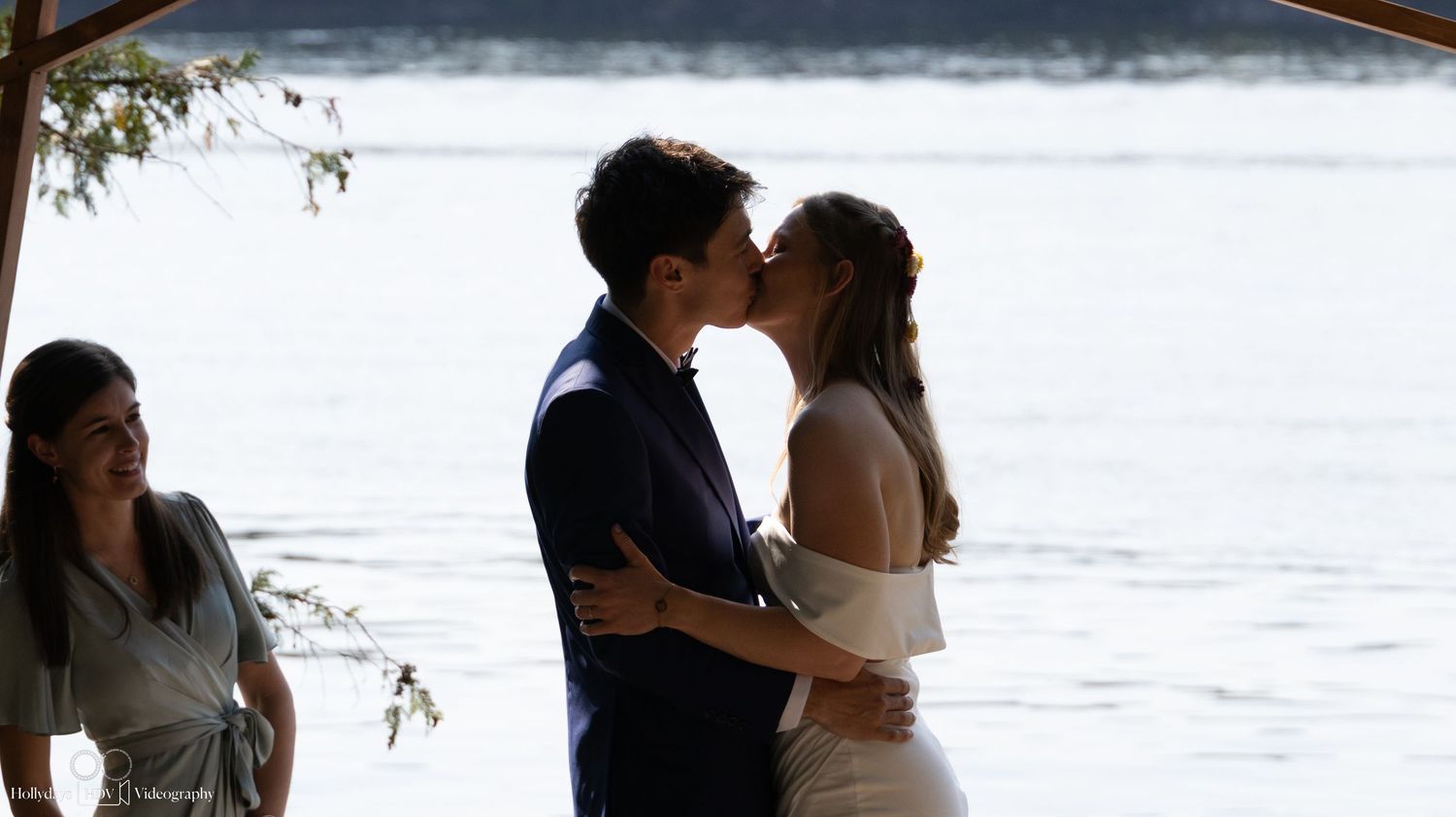 A couple shares a romantic kiss during their lakeside wedding ceremony as guests look on.