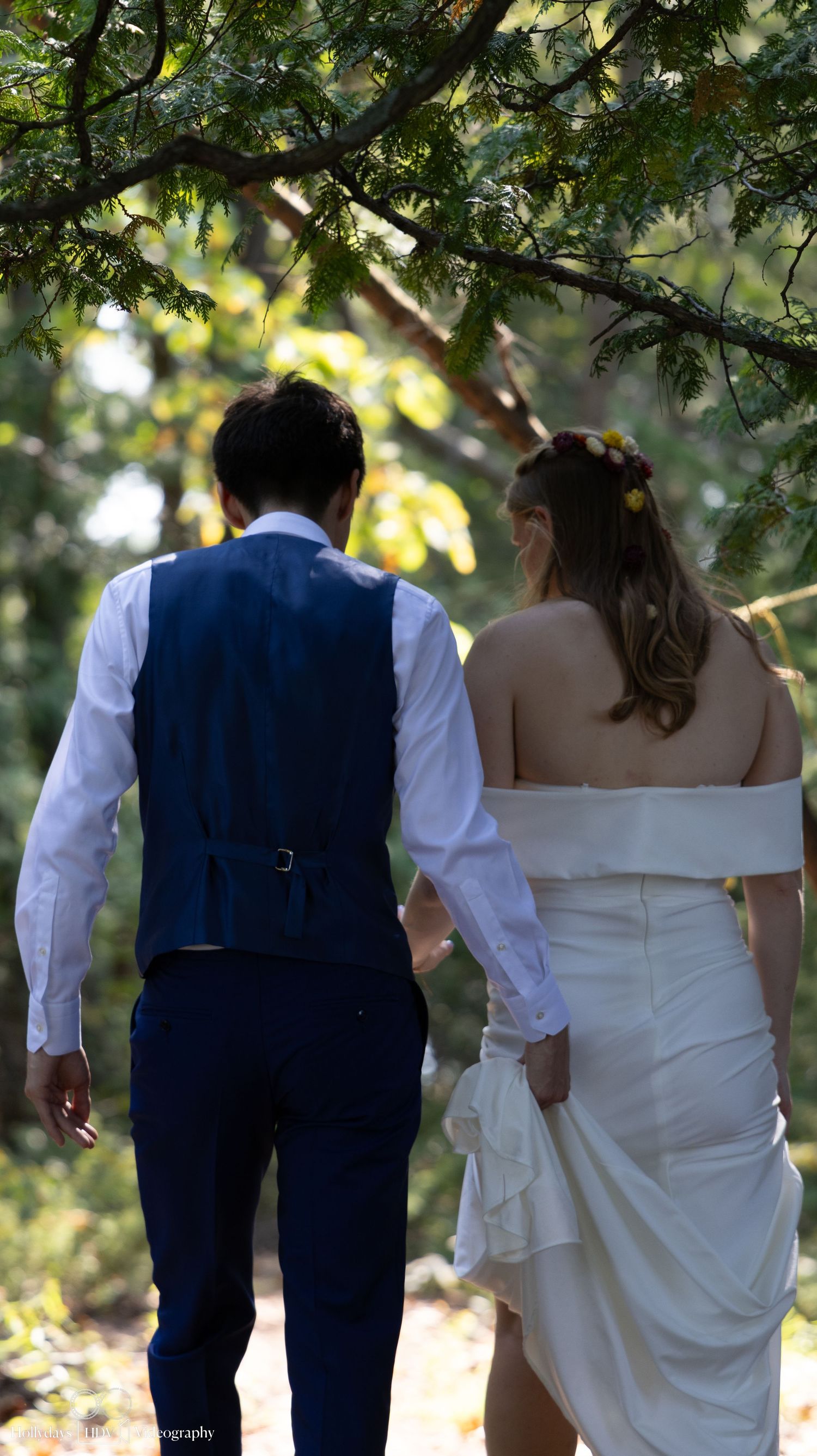 Couple in formal attire walking together under tree canopy.