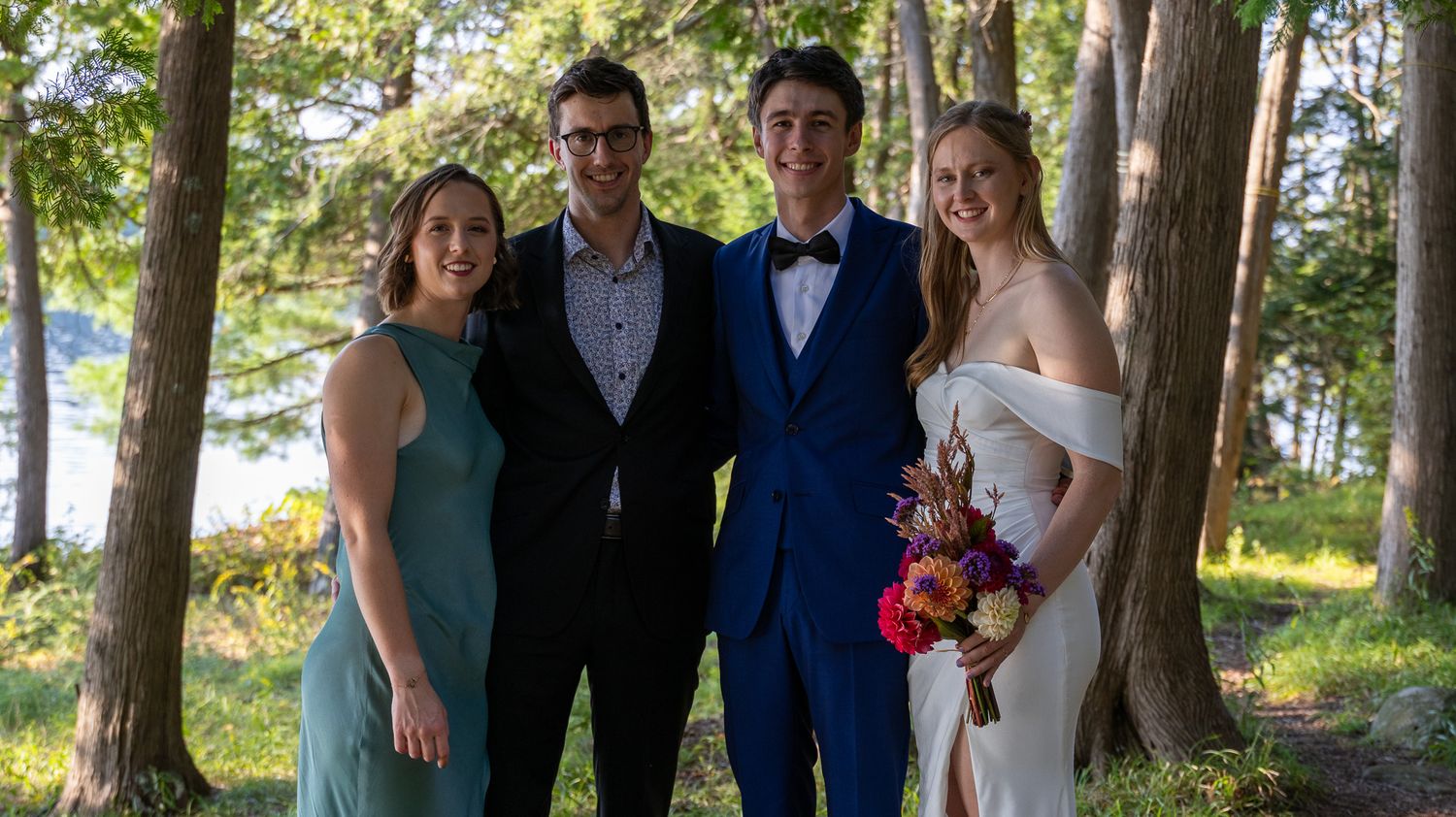 Wedding party poses together in formal attire among trees by lakeside.