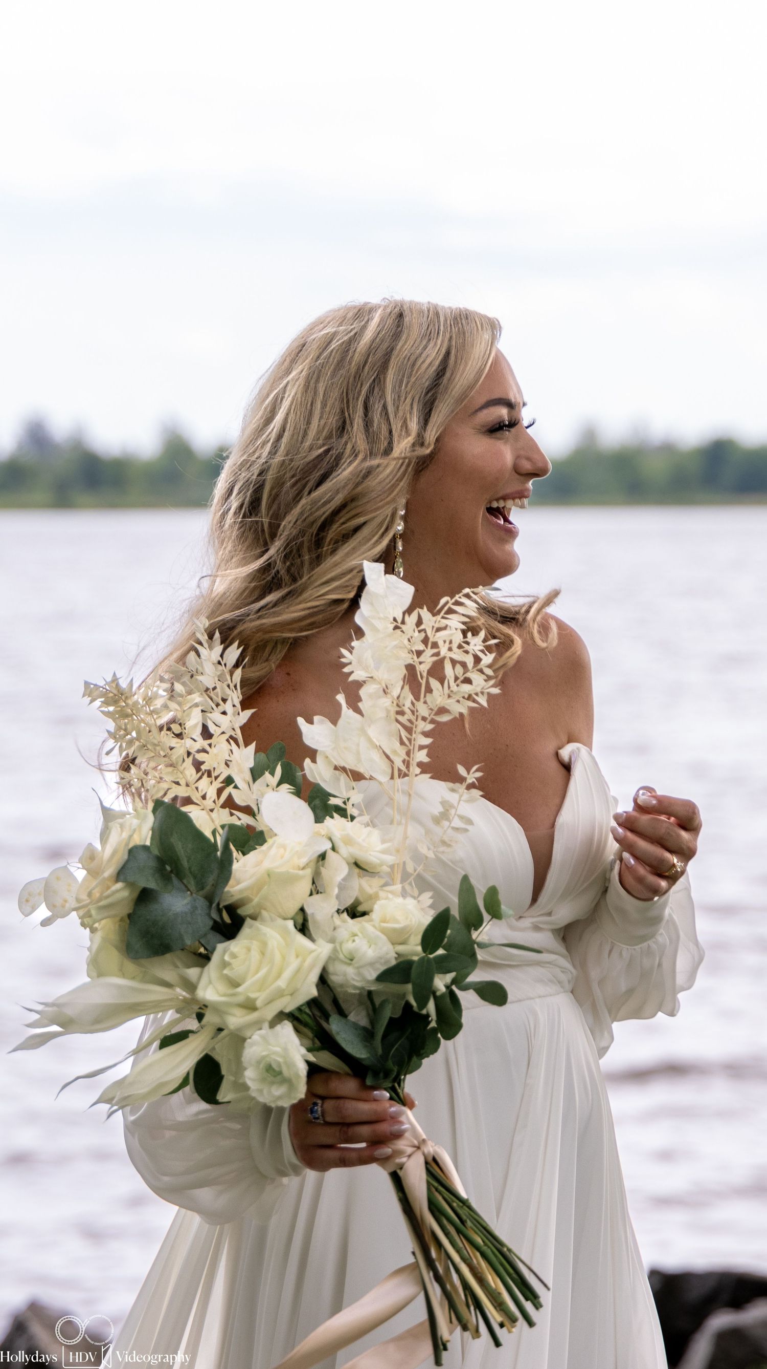 Joyful moment captured at waterfront wedding with flowing white dress and elegant bouquet of cream roses and greenery.