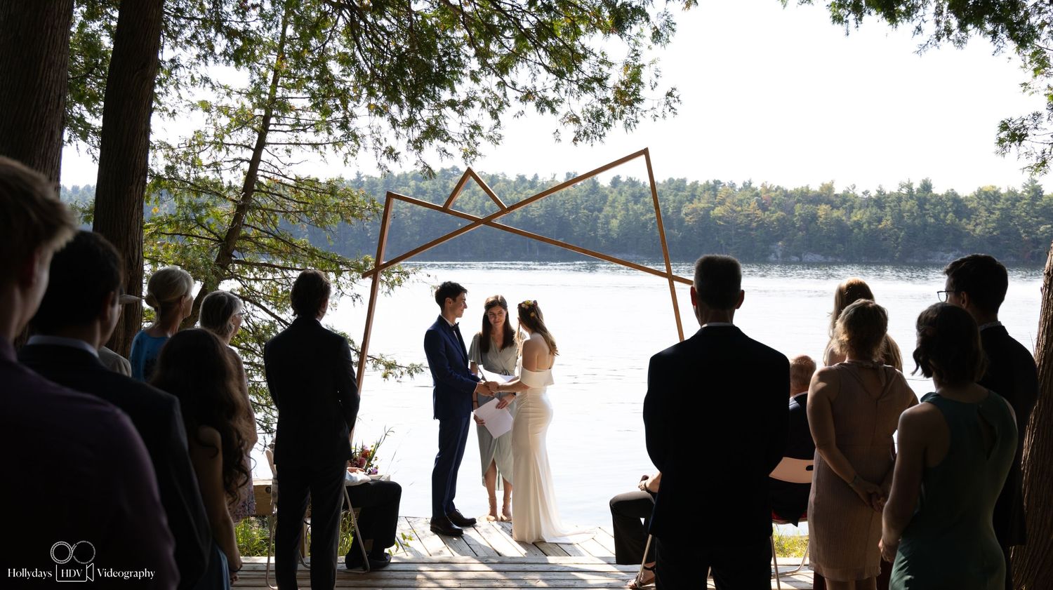 A silhouetted wedding ceremony by a lake with guests watching the couple exchange vows under wooden beams.