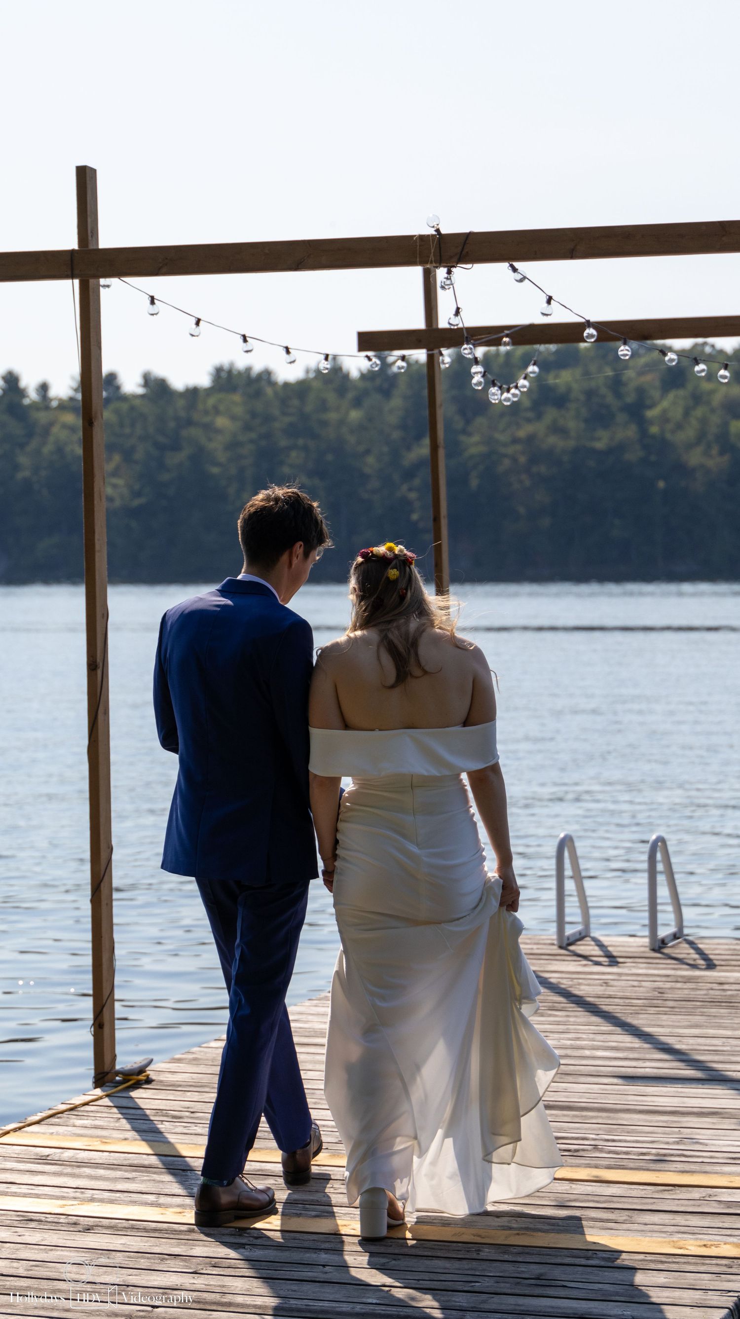 A couple in wedding attire stands on a lakeside dock with strung lights overhead during a sunset.