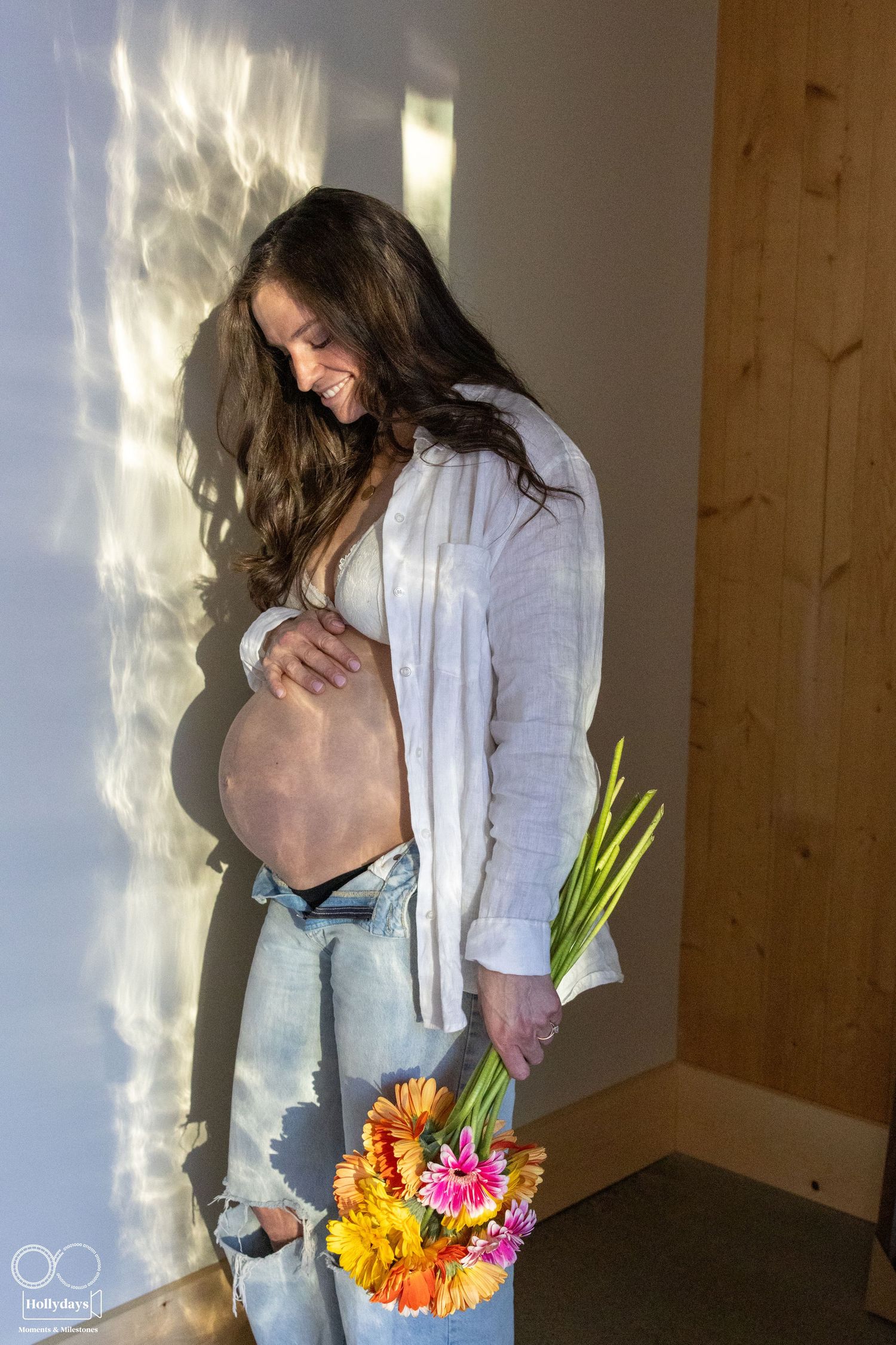 Expectant mother in casual attire holds her belly while standing by window with colorful spring flowers. Maternity photography