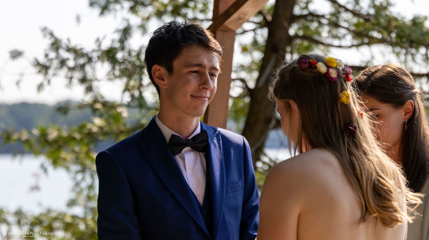 A wedding ceremony taking place outdoors with a groom in navy suit and bow tie smiling during the service.