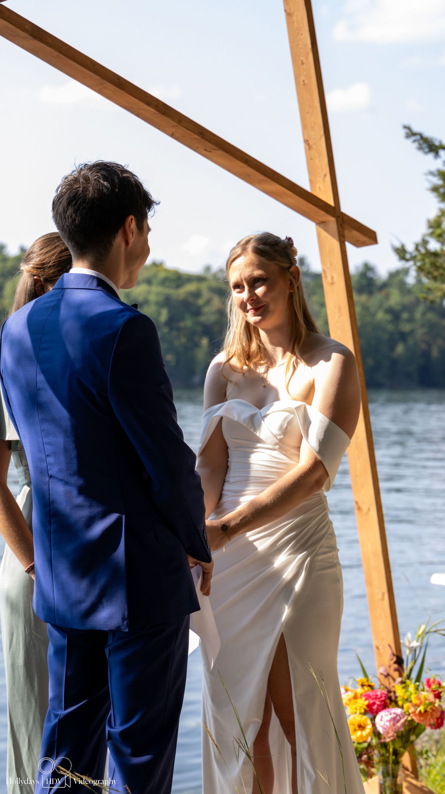 A couple exchanges vows at a waterfront ceremony under a wooden arch with colorful floral arrangements.