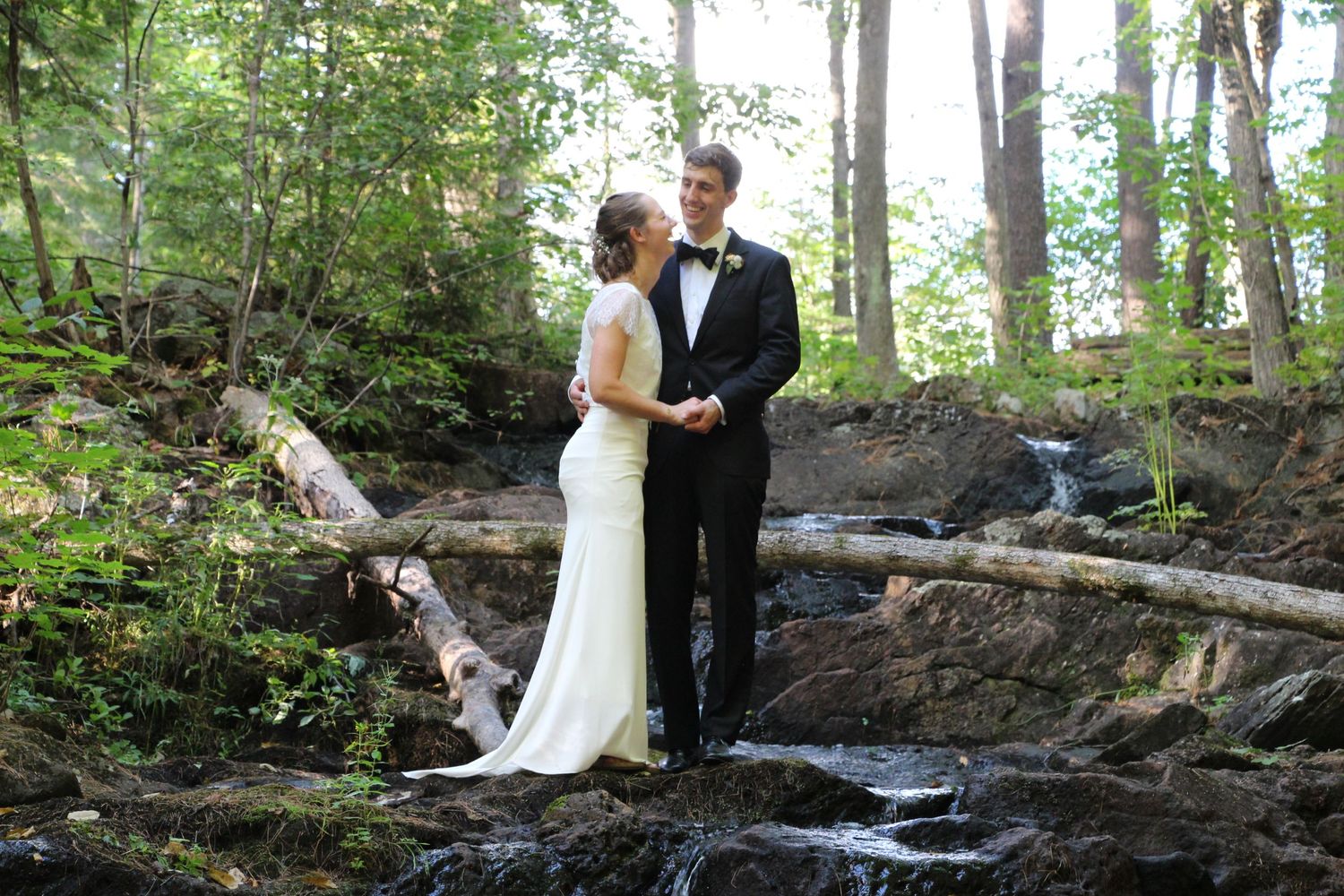 A couple shares an intimate moment in their wedding attire while standing on rocky terrain in a lush forest setting.