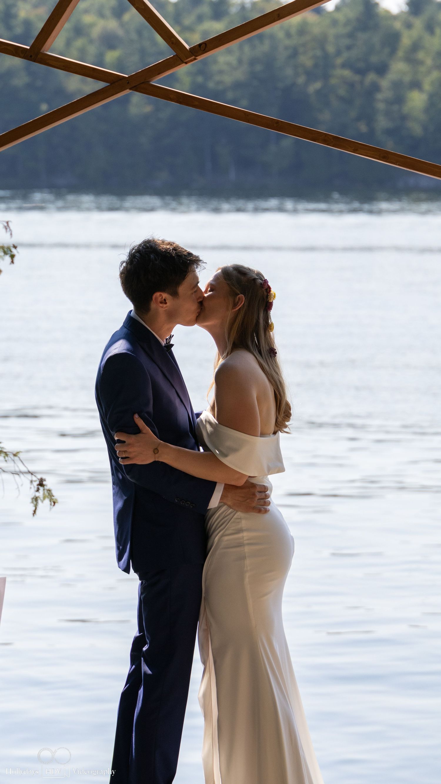 A couple shares their first kiss during an outdoor lakeside wedding ceremony under wooden beams.